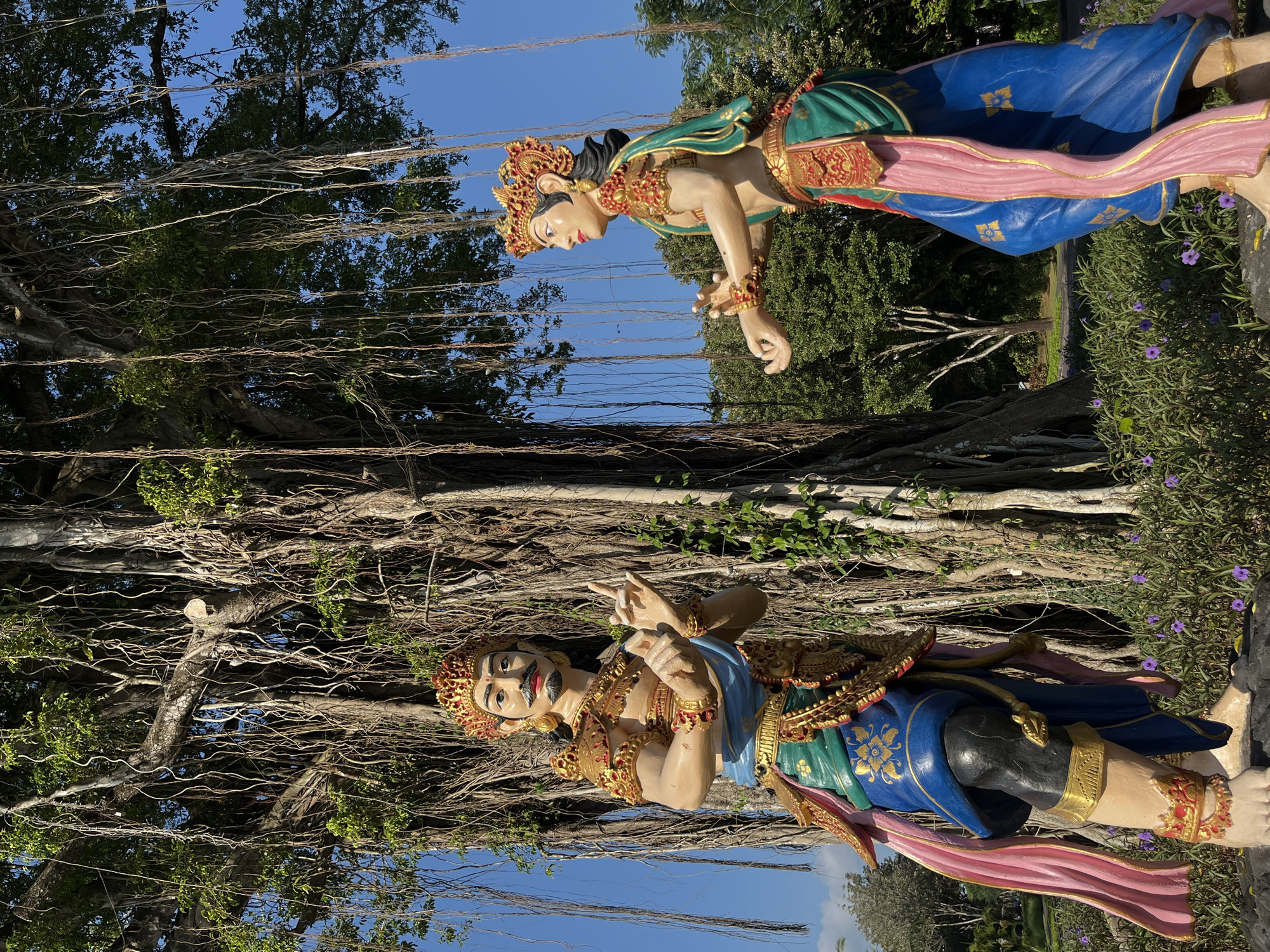 Two statues of Balinese dancers, a male and a female, stand beneath a large banyan tree with its characteristic aerial roots. The statues are adorned in traditional Balinese attire, featuring ornate crowns, gold-colored jewelry, and vibrantly colored fabrics in blue, green, pink, and red. The male statue, on the left, has a mustache and a stern expression, with his hands positioned as if in a gesture of storytelling or instruction. The female statue, on the right, faces to her left, with her hands also in a graceful dance pose. The banyan tree's massive trunk and cascading roots dominate the background, with lush greenery and a clear blue sky visible. The statues are positioned on dark, rocky pedestals surrounded by a bed of purple flowers and green foliage. This scene is located in Kuta Selatan, Indonesia.