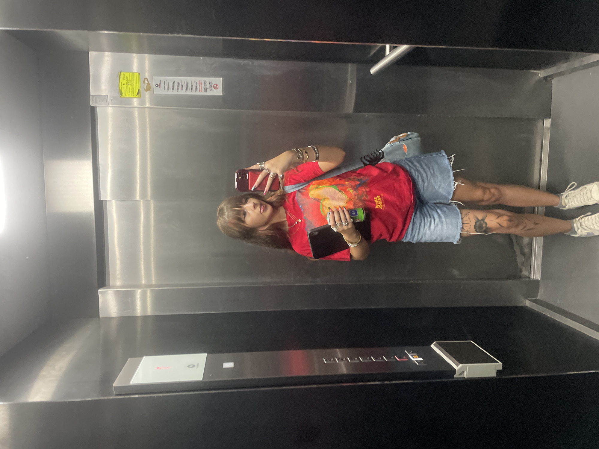 A young woman with bangs and shoulder-length brown hair with reddish highlights takes a mirror selfie in an elevator. She is wearing a red t-shirt with a graphic print, denim shorts, and white sneakers. She is holding a tablet in her left arm and a green can in her right hand, and her tongue is sticking out. She is also wearing several rings and bracelets. The elevator is made of stainless steel with black panels. There are buttons on the left wall of the elevator. The image was taken in Raanana, Israel.