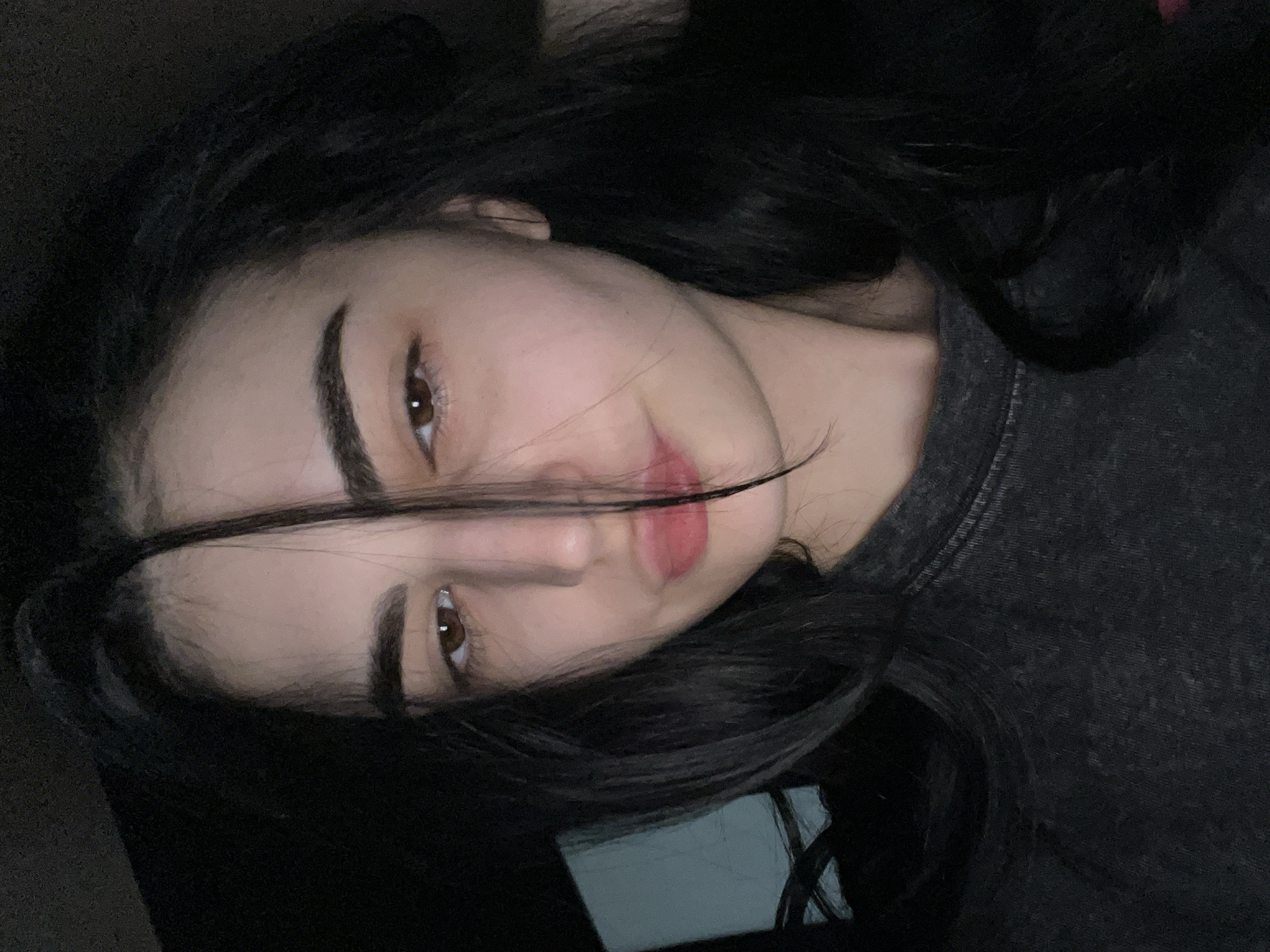 A close-up photograph depicts a young woman with dark hair and brown eyes. A strand of her hair falls across her face. Her eyebrows are thick and dark. She has light skin, and her lips are slightly parted, revealing pink color. She is wearing a dark gray shirt. The background is dark and indistinct. There is no indication of the location being Burgas, Bulgaria.