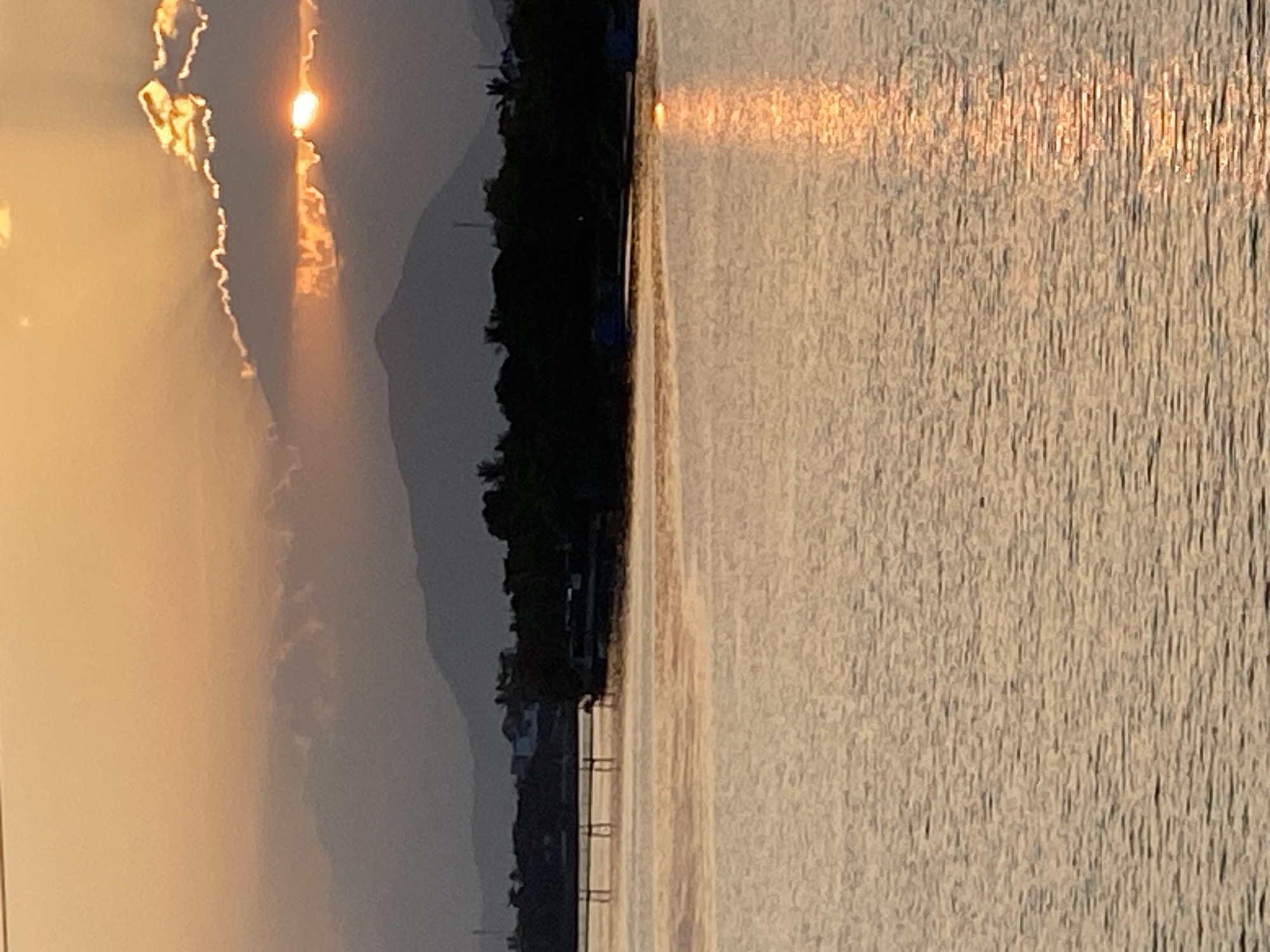 The image captures a sunset over the Cai River in Nha Trang, Vietnam. The sun is partially obscured by clouds, casting a warm orange glow across the sky. The river's surface reflects the sunset, creating a shimmering path of light. In the foreground, the calm water dominates the lower half of the frame. The middle ground reveals a treeline along the riverbank, with hints of buildings and structures suggesting a populated area. A pier or jetty extends into the water from the left. The background features rolling hills or mountains, fading into the hazy distance under the twilight sky. The overall scene is serene and natural, emphasizing the beauty of the sunset over the waterway.