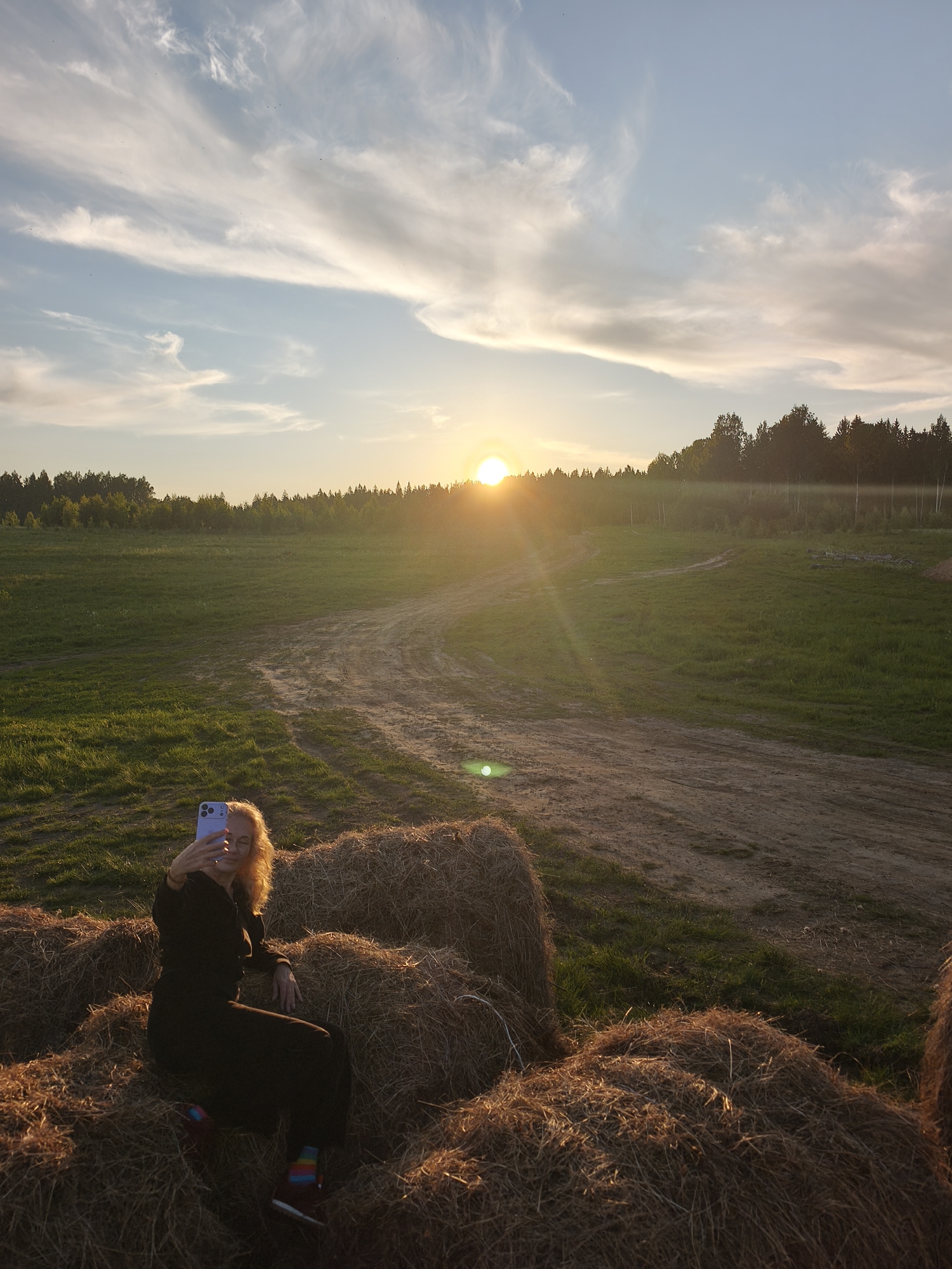 The image captures a serene rural landscape at sunset, located in Стеклянный, Russia. In the foreground, a woman with curly, light-colored hair is seated comfortably on large, stacked hay bales. She is dressed in dark, long-sleeved attire and dark pants, with visible colorful striped socks. She holds a light purple smartphone in her right hand, taking a selfie, with the device angled slightly towards her face.
The setting is an expansive open field covered in green grass, with a winding dirt path snaking through it towards the background. A dense line of trees, forming a forest, stretches across the horizon, appearing somewhat silhouetted against the sky. The sun is a prominent feature, low on the horizon on the right side of the frame, casting a bright, golden glow and warm light across the scene, indicative of either sunrise or sunset, most likely sunset given the warm hues. Wispy, elongated clouds are scattered across the light blue and orange-tinged sky. The overall atmosphere is peaceful and warm, highlighting the beauty of the natural environment during the golden hour. There is no visible text in the image.