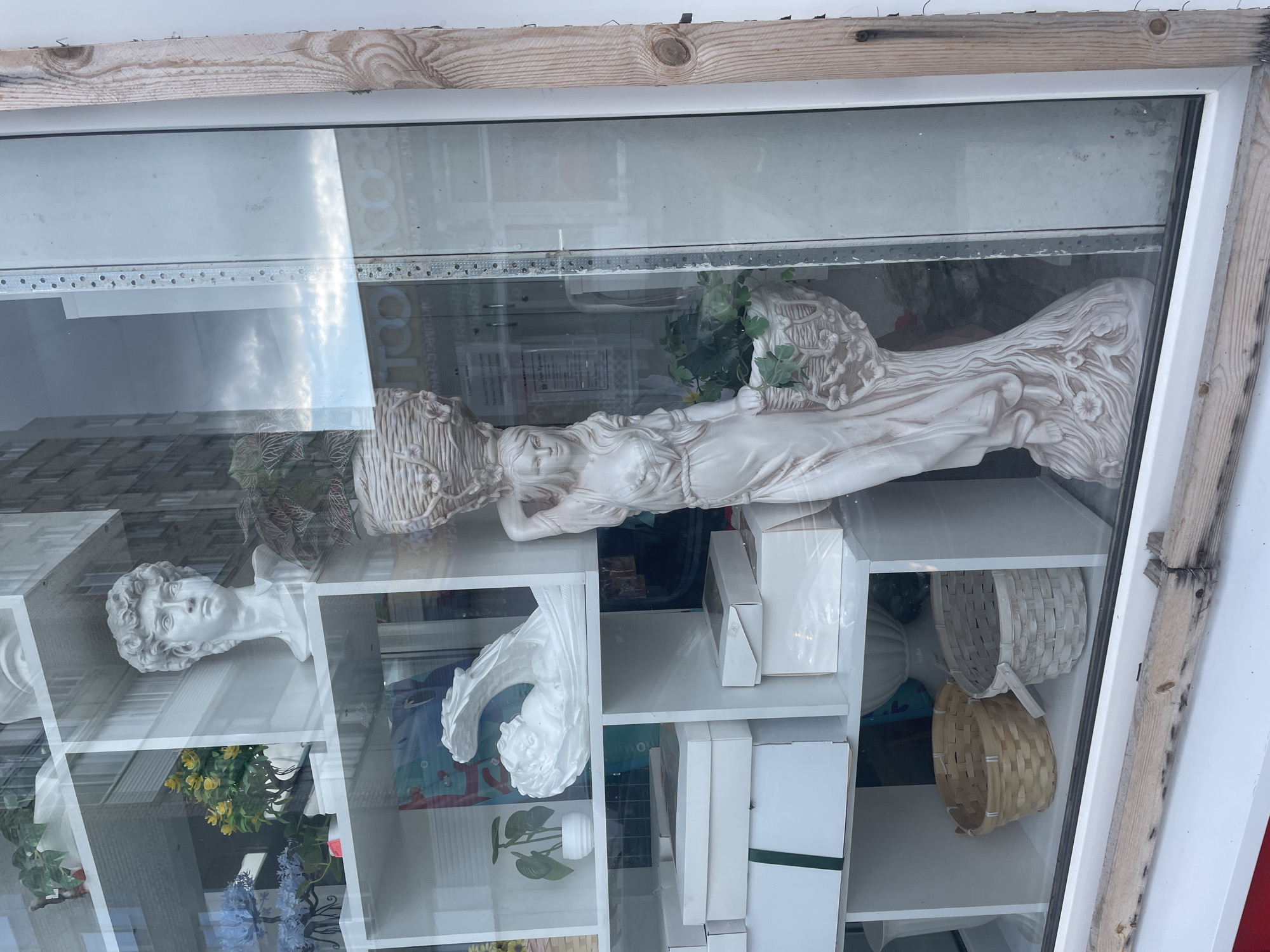 A retail display window in Chaikovsky, Russia, showcases decorative items. The window is divided into several shelves, each filled with various objects. Prominently featured is a large, pale statue of a woman carrying a basket of plants on her head. To the left, on a higher shelf, is a bust of a man, possibly a replica of Michelangelo's David, alongside artificial blue flowers and a potted plant. Below these, white boxes are stacked, some tied with green ribbon. Further down, two woven baskets are visible. The display is illuminated, and reflections of a building and the sky are visible on the glass. The exterior frame of the window includes weathered wood.