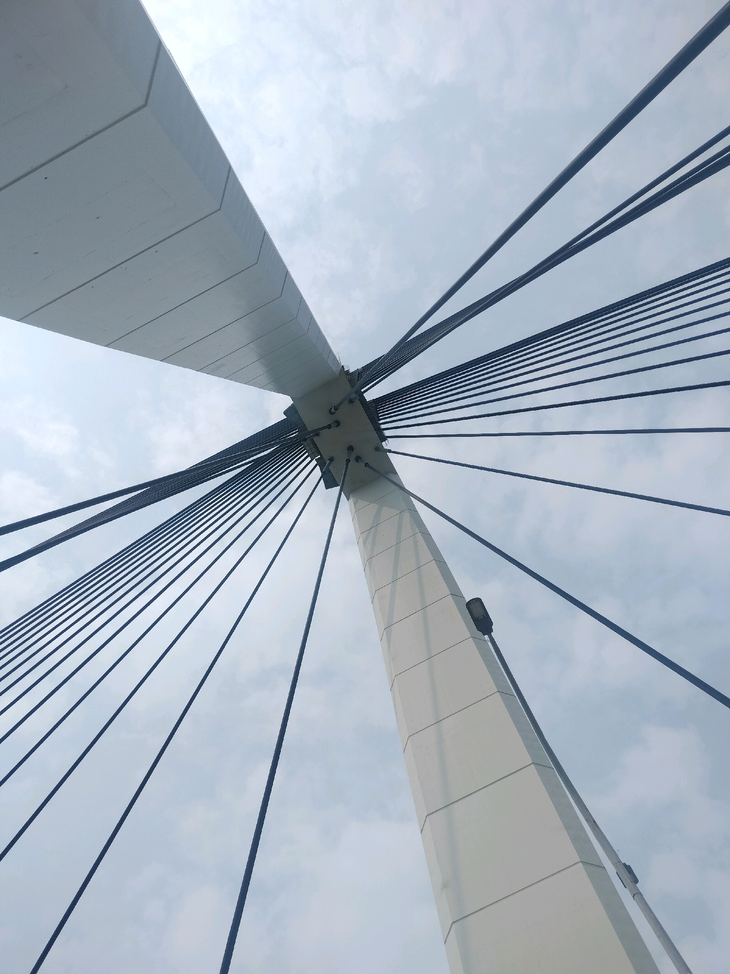 The image is a low-angle shot of a modern bridge structure against a cloudy sky. The main subjects are a large, white, angular pylon and numerous dark cables that fan out from the top of the pylon towards the viewer's right. The pylon is segmented with visible panel lines and a small, dark light fixture is attached to its side. A broad, white beam of the bridge extends diagonally from the upper left of the frame. The perspective emphasizes the scale and geometry of the bridge's engineering. This perspective is consistent with viewing the Batam cable-stayed bridge from below.