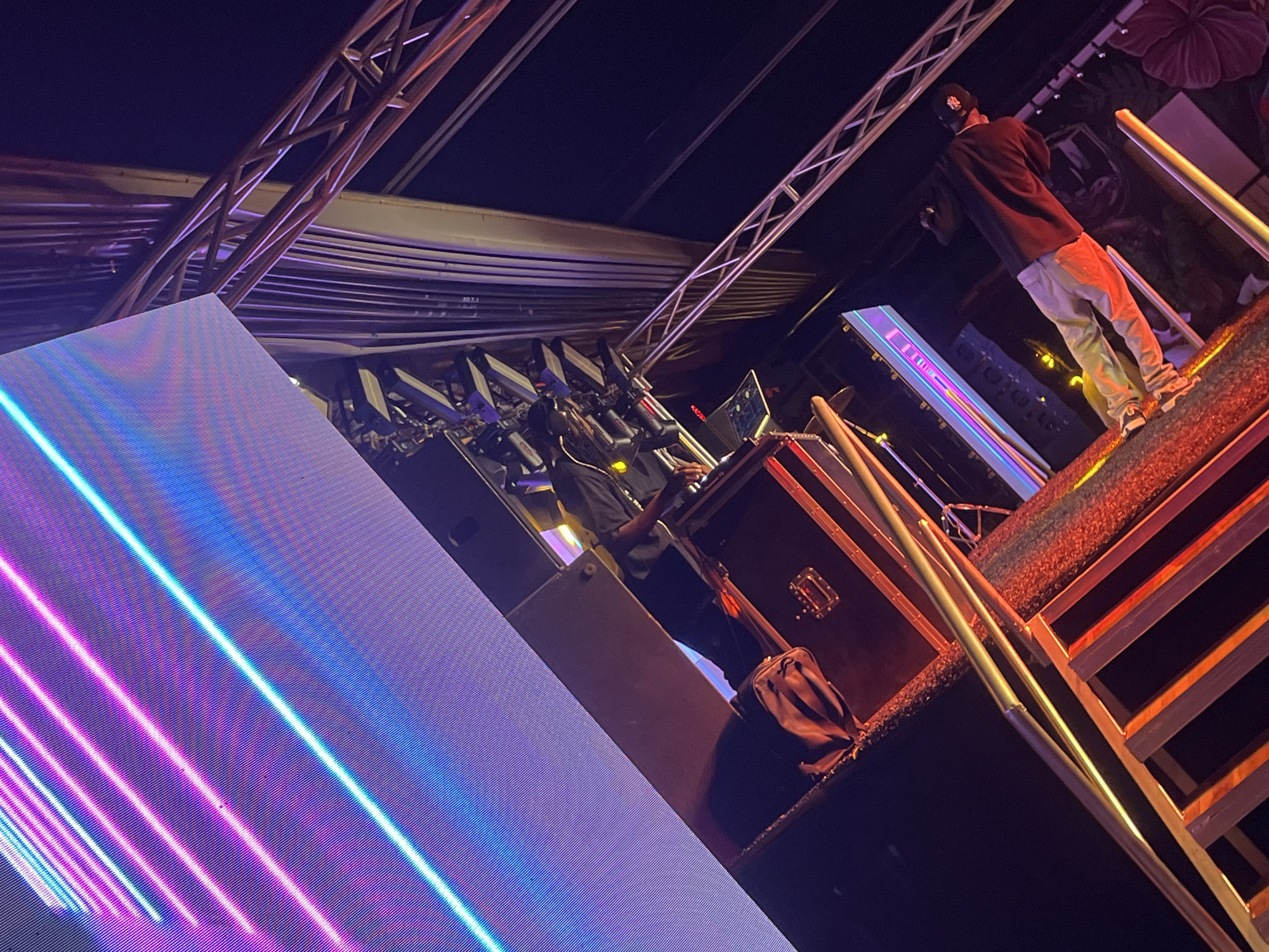 A stage setup is captured indoors, possibly at a concert in Howayi, Nigeria. Two performers occupy the platform. One is operating electronic equipment, likely a DJ set. The second is standing near the platform's edge, wearing a dark long-sleeved shirt, light trousers, and a baseball cap. Stage lights and a large LED screen projecting linear patterns dominate the left part of the visual space. Structural scaffolding is visible above.