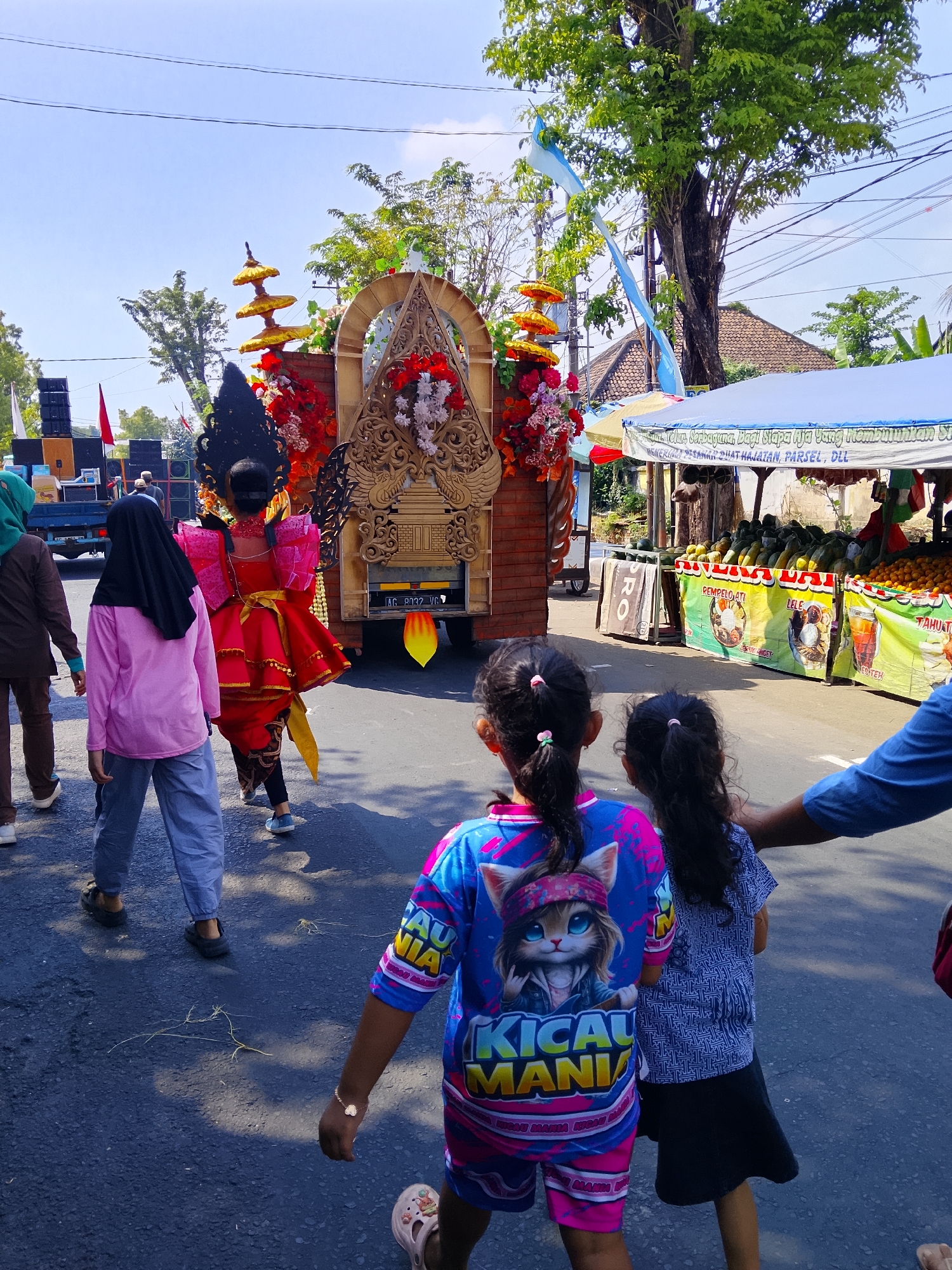 The image depicts a street scene in Kertosono, Indonesia, likely during a cultural event or parade. In the foreground, two young girls are walking away from the camera. The girl on the left wears a vibrant blue and pink t-shirt featuring a cartoon cat and the text "KICAU MANIA". The girl on the right wears a blue patterned shirt and a dark skirt. A person's arm, wearing a blue shirt, is visible on the right side of the frame, with their hand resting on the girl on the right's shoulder.
Behind the children, a person in a striking red and gold traditional costume, adorned with a large black headdress, is walking. Further back, a decorated float is visible, mounted on a truck. The float is adorned with red and white flowers, intricate golden carvings, and golden tiered ornaments. Loudspeakers are positioned on the left side of the image, suggesting music or announcements. In the background, market stalls are set up under white awnings, displaying various fruits and produce. The scene takes place on a paved road under a clear blue sky with green trees lining the sides.