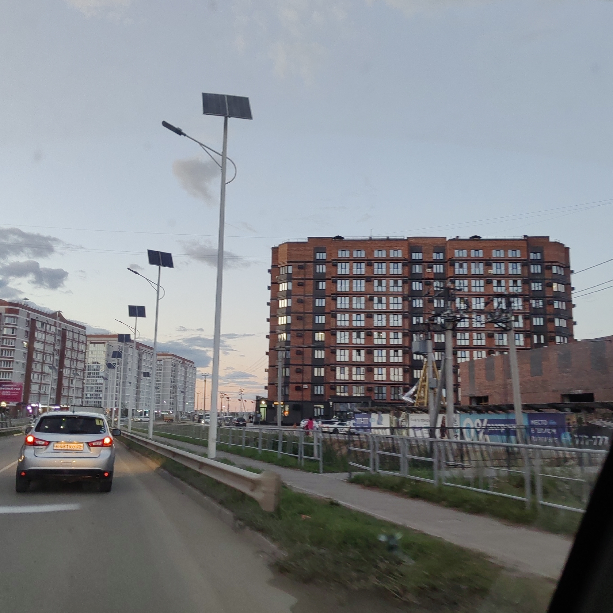 The image captures a street scene in Chigiri, Russia, likely during the evening or early morning given the soft, diffused light and the illuminated windows of the buildings. The foreground features a silver hatchback car driving on a paved road. The car has its brake lights illuminated and a license plate with Cyrillic characters and numbers that appear to read "H481KO 28". To the left of the road, a concrete barrier with a metal railing separates the road from a pedestrian walkway and grassy area.
Dominating the background are several multi-story apartment buildings. The building closest to the right is constructed with dark red brick and features numerous windows with white frames, some of which are lit from within. To the left of this building are other apartment complexes, appearing lighter in color and also featuring many windows. Several streetlights, equipped with solar panels, are visible along the roadside. In the mid-ground, a construction site is apparent with scaffolding and unfinished brick structures. The sky above is overcast with scattered clouds.