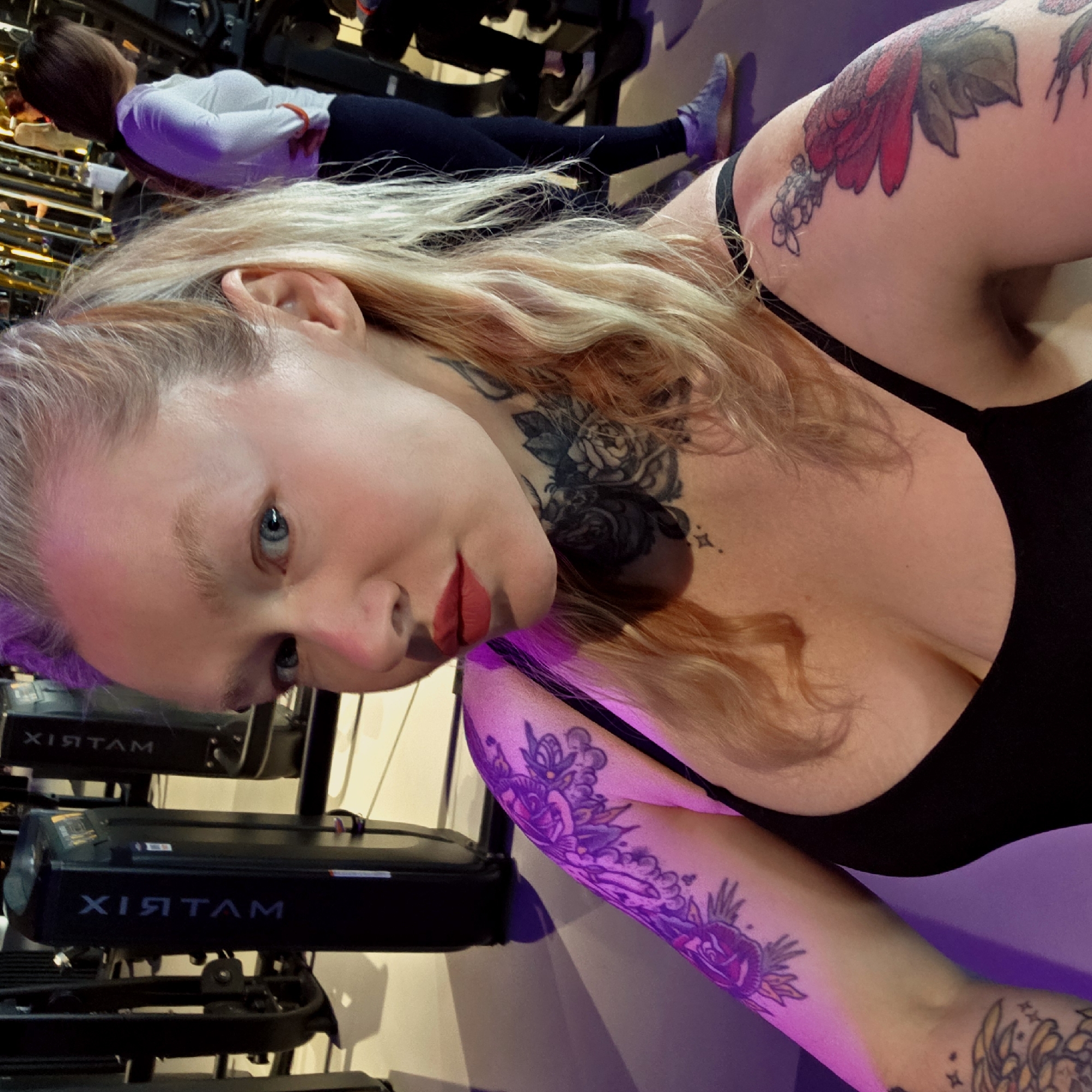 This image is a selfie-style shot taken in what appears to be a modern gym or fitness studio in Saint Petersburg, Russia.
The main subject is a woman with long, wavy blonde hair, fair skin, and striking blue eyes. She is looking directly at the camera with a neutral expression, enhanced by red lipstick. She is wearing a black bra or tank top with thin straps. Her arms and upper chest are adorned with intricate tattoos; a prominent black and grey rose design covers her neck/upper chest area. On her left shoulder/upper arm, there is a large, colorful tattoo featuring red flowers and green leaves, while her right forearm displays vivid purple and yellow floral patterns.
In the background, another woman with brown hair is visible, wearing a white long-sleeved top and dark leggings. She appears to be exercising or stretching, with her legs extended, possibly on a piece of gym equipment. The setting features multiple pieces of dark exercise equipment. One machine clearly shows the brand name "MATRIX" (visible as "XIRTAM" due to the camera angle) in blue lettering. The lighting in the space has a noticeable purple and pink hue, suggesting artificial colored lights. The overall scene depicts a moment within a workout environment.