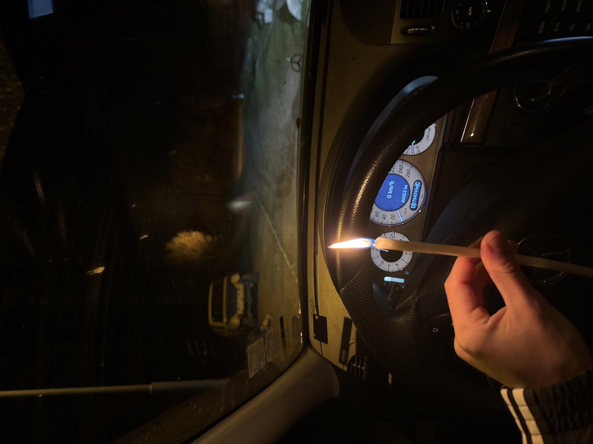 From the interior of a Mercedes-Benz, a hand holds a lit stick near the instrument panel. The odometer reads 6602.2 km and the car is stationary (0 km/h). Outside the car window, the exterior environment is dimly lit, showing what appears to be a road and possibly other vehicles. The image was taken at Buhovo, Bulgaria as indicated by the plate number which follows the Bulgarian numbering scheme.