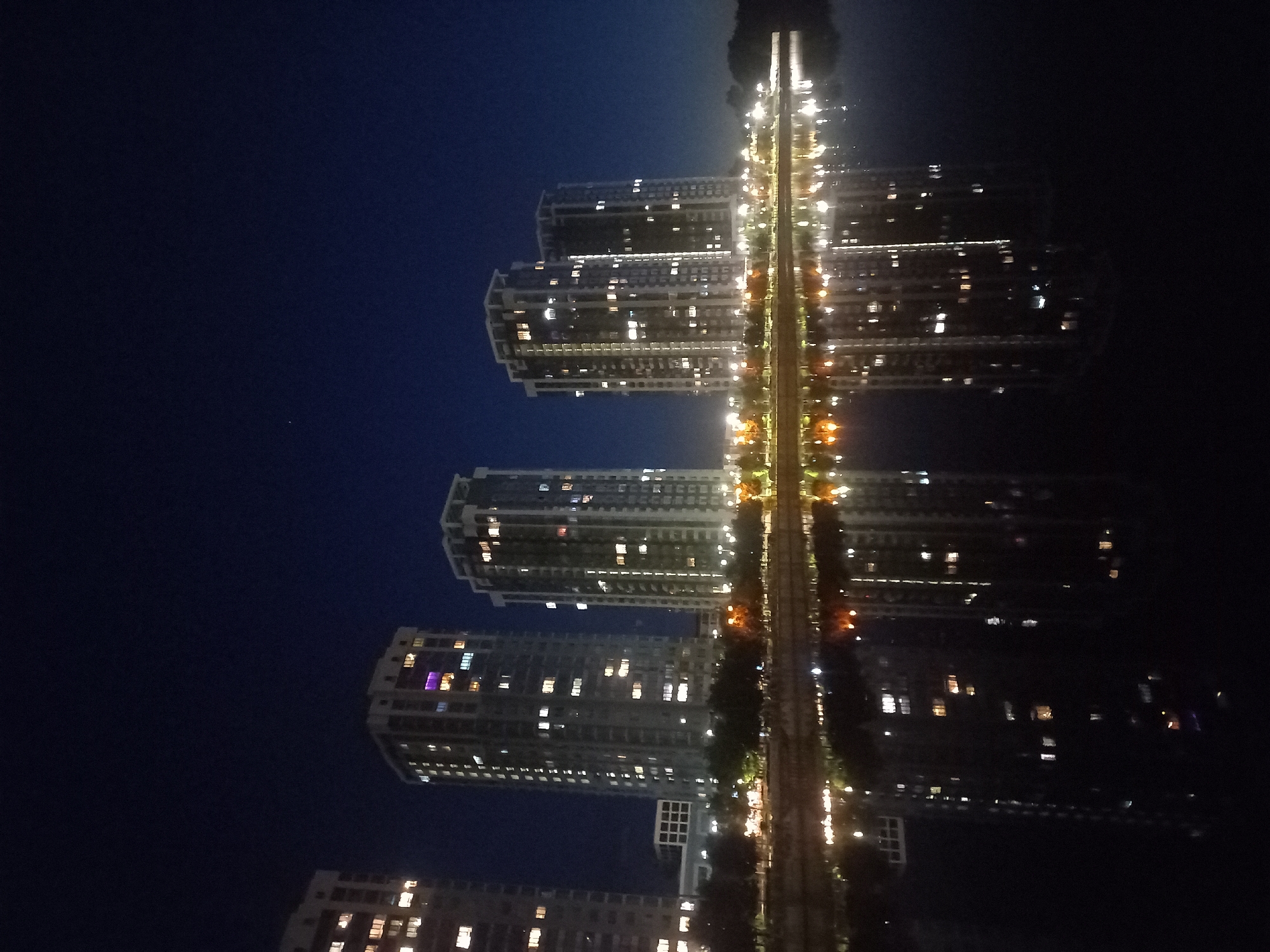 At night, a line of buildings and their reflections line a strip of light. The buildings are situated on the left side of the image, and the reflections are on the right. The sky appears dark blue. The lights from inside the buildings shine through the windows. This scene is located in Kyiv, Ukraine.