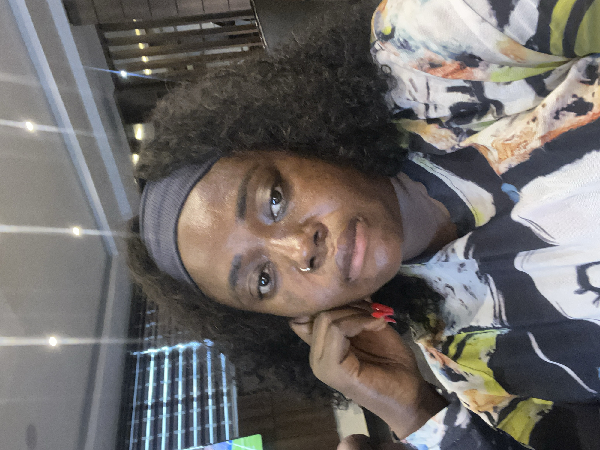 A medium shot captures a woman with dark skin and curly hair from the chest up, looking directly at the camera. She is wearing a grey headband and a multi-colored patterned shirt. Her left hand is raised, with her fingers touching her ear and her bright red-painted fingernails visible. A small silver hoop is visible in her nostril. The background appears to be an indoor setting, possibly a restaurant or lounge, with blurred elements of wooden paneling and overhead lighting. The overall lighting suggests a dimly lit environment. The scene is likely in Abuja, Nigeria.