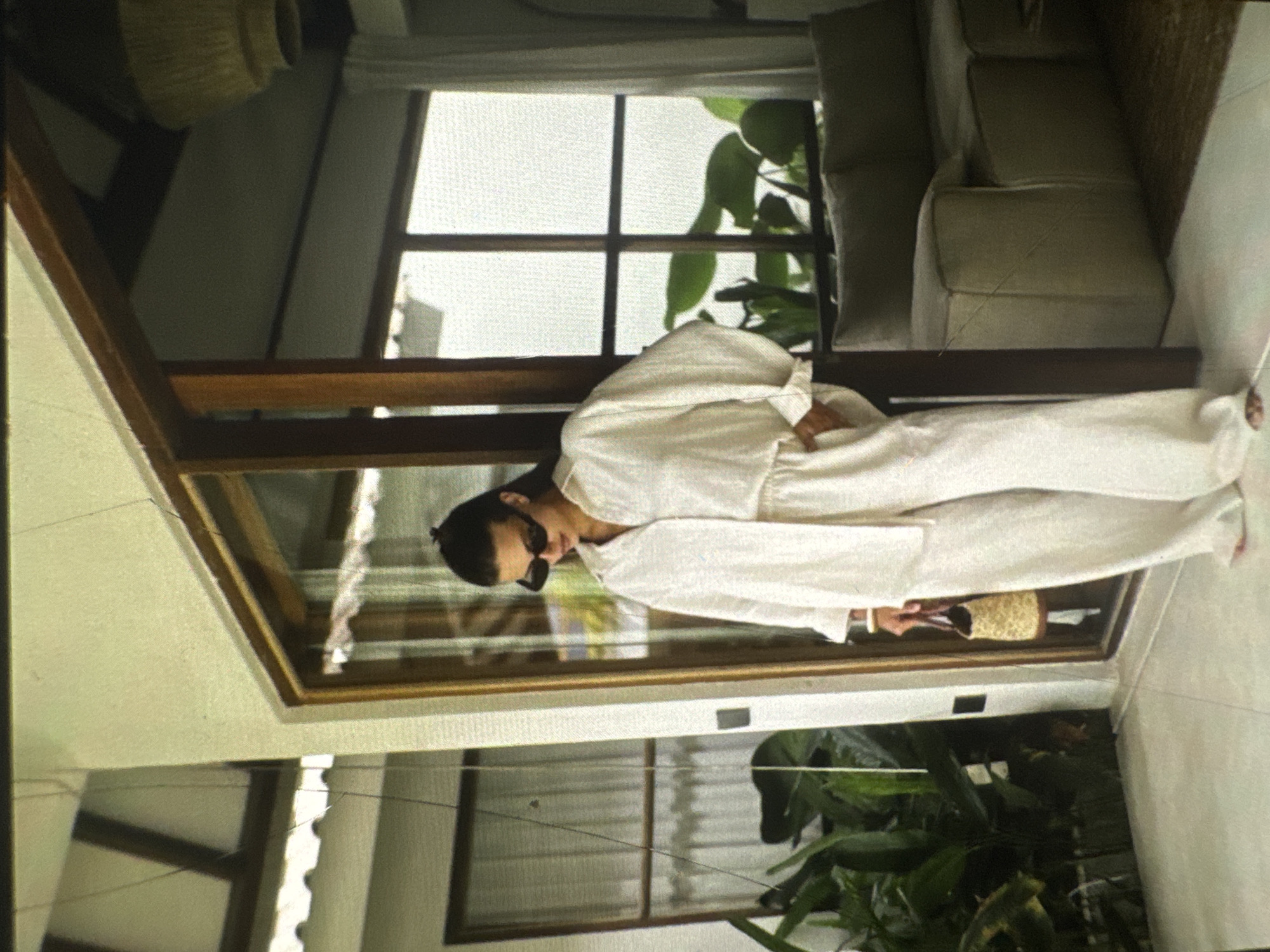 The image shows a woman standing indoors, possibly in a house or a cafe, wearing a white linen shirt and matching pants. She is also wearing sunglasses and holding a straw bag. The environment appears to be modern and minimalist, with large windows offering a view of lush greenery. The woman is looking down, her hands in her pockets. The scene is well-lit, with natural light coming through the windows. There is a sofa visible in the background, suggesting a living area. The location is specified as Protasovo, Russia.