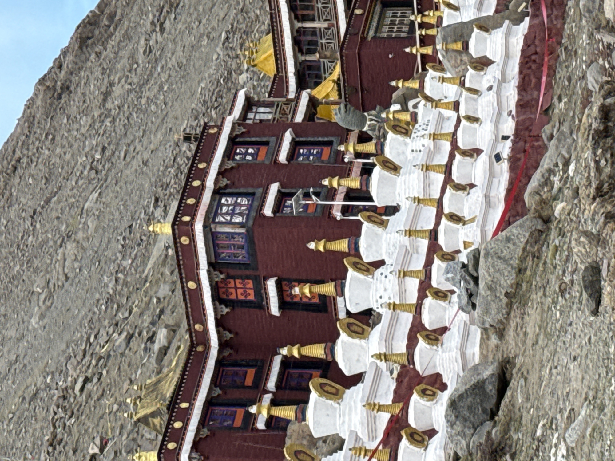 The image shows a temple or monastery built into the side of a mountain. The building has multiple levels, with a facade of maroon walls, white trim, and numerous windows with colorful frames. Adjacent to the building, and partially obscuring it, is a row of white structures resembling stupas, each with a golden or brass-colored finial. The mountain is composed of gray, rocky terrain. The scene appears to be set in a remote and rugged location. It is unlikely to be Parga town, China.