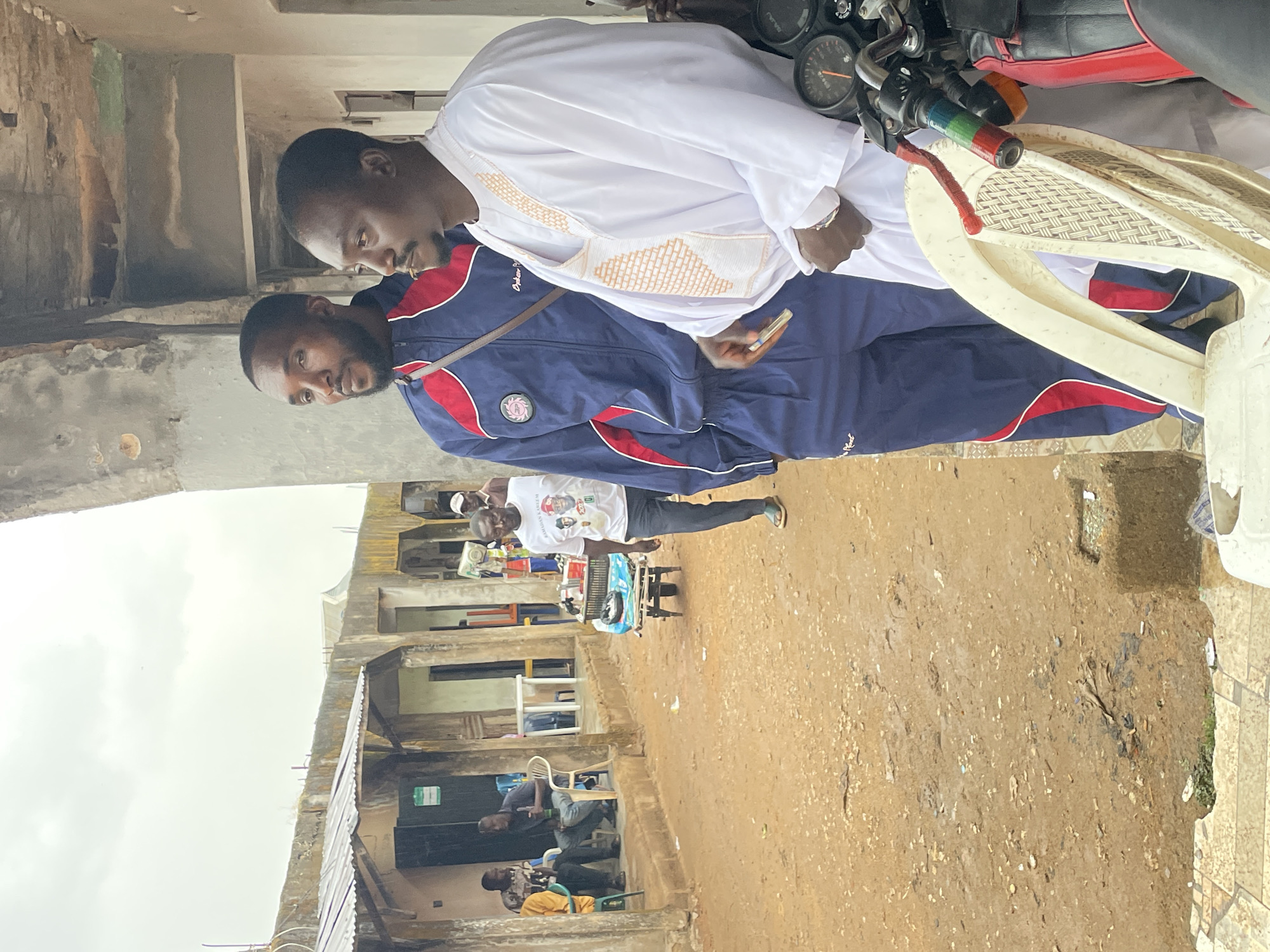 Two African men are seated on a parked motorcycle. The man to the right is wearing a white robe, while the other man is wearing a blue and red tracksuit. The man in the tracksuit has a strap across his chest. The motorcycle has a black seat with a red stripe. The motorcycle handlebars have rainbow-colored grips. In the background, a building under construction can be seen. Four men are sitting in front of the building. Another man is pushing a cart behind the men in front of the building. The ground is bare earth. The location is Jos, Nigeria.