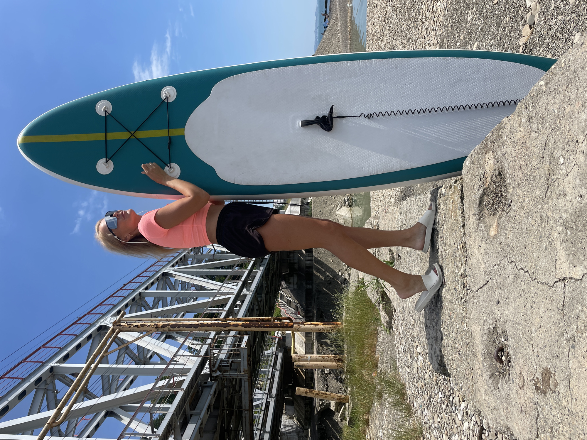 A blonde woman stands on a concrete and pebble-covered embankment, holding a turquoise and white stand-up paddleboard. She is wearing a pink crop top, black shorts, white slides, and sunglasses, with earbuds in her ears. The paddleboard is upright, leaning against her. In the background, a large, industrial-looking metal bridge with red railings spans across the water. The sky is clear blue with a few scattered clouds. The environment appears to be coastal or near a body of water, with a rocky shore and some sparse vegetation.