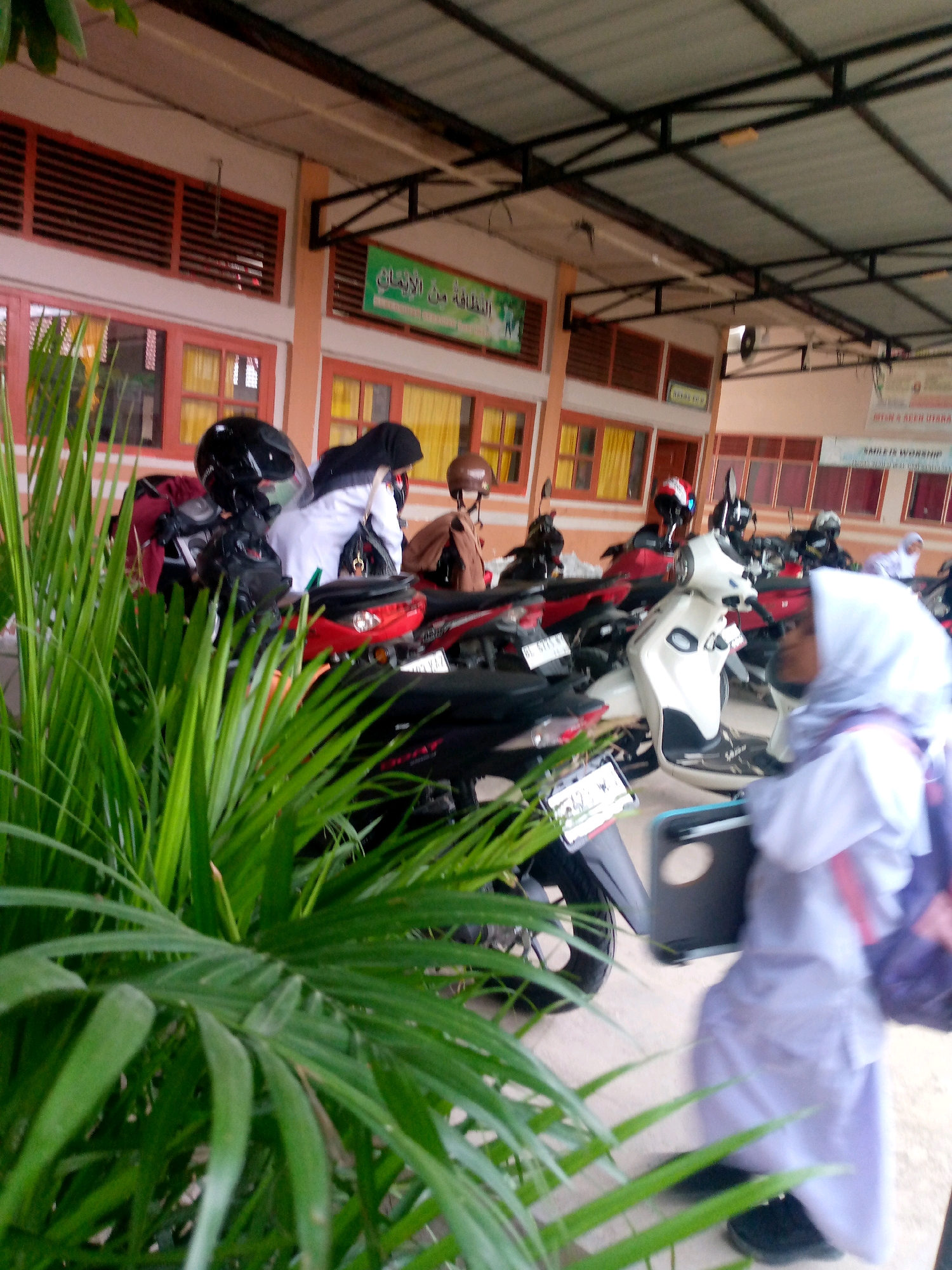 In an outdoor, covered area that appears to be a school or religious institution, several parked motorcycles are visible. A few individuals, likely students, are present. One person in the foreground is wearing a light purple hijab and a white uniform, carrying a purple backpack. Another person in the background is wearing a black hijab and a white garment. The environment is characterized by a building with orange-toned walls and windows with orange frames. Green foliage is prominent in the foreground, suggesting a tropical or subtropical setting. A green sign with Arabic script is mounted on the building wall. The ground is paved. The lighting suggests daytime.