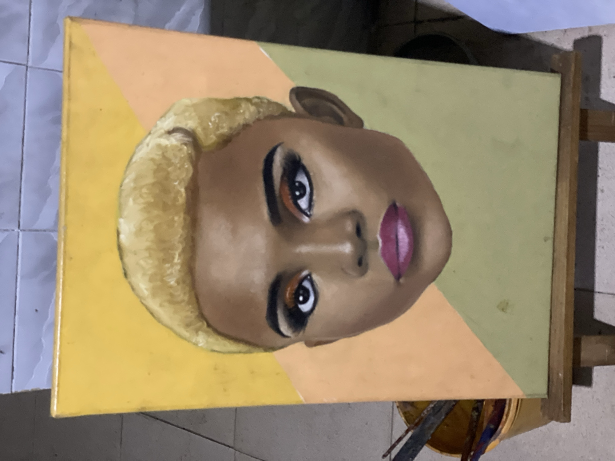 An oil painting of a Black woman with blonde hair is displayed on an easel indoors. The background of the painting consists of geometric sections colored yellow, peach, and light green. The painting is likely located in Ado, Nigeria.