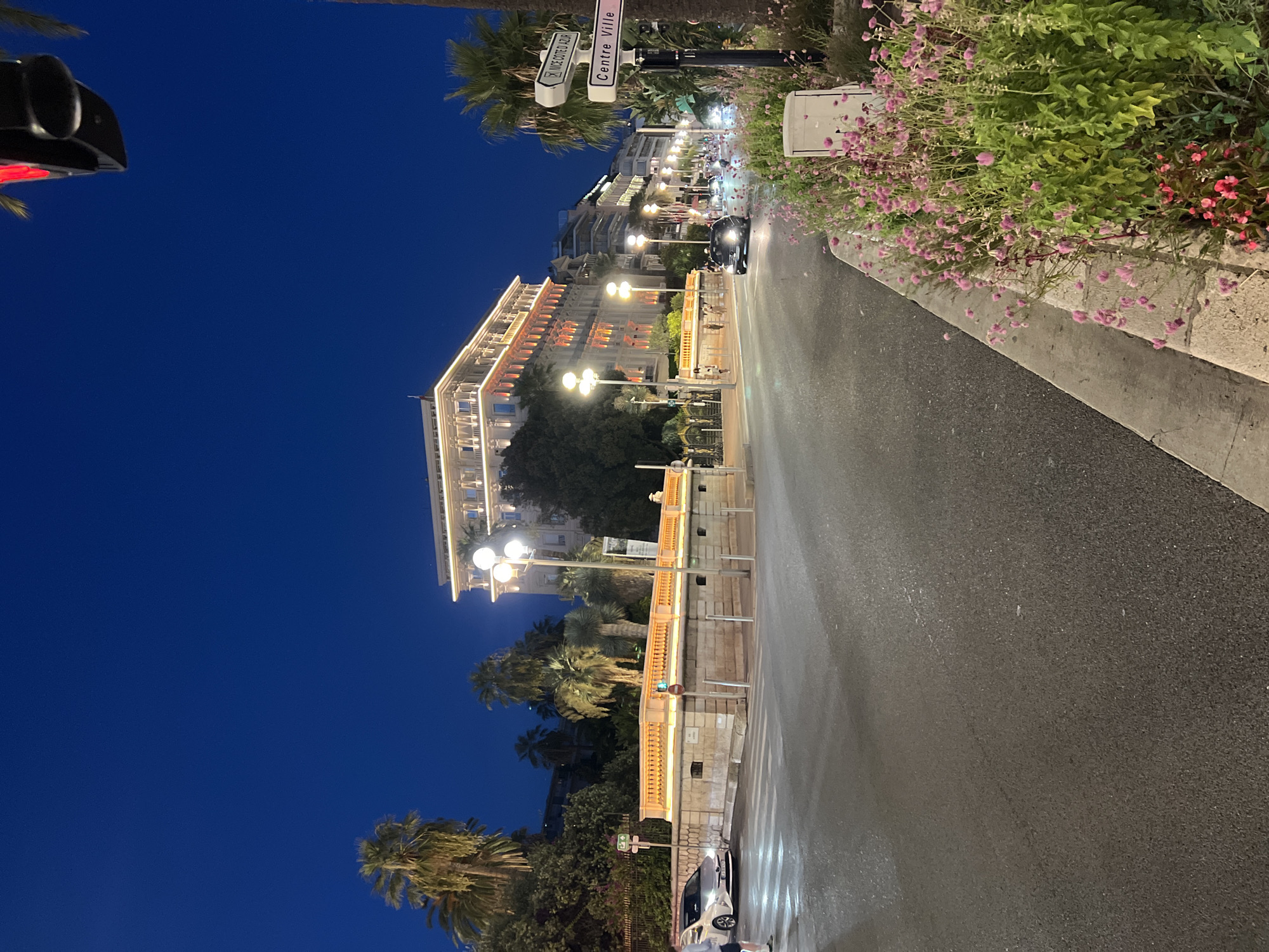 This image depicts a street scene in Nice, France, at twilight. In the background, a large, ornate building, likely a hotel, is illuminated with warm lights. Palm trees are visible throughout the scene, characteristic of the Mediterranean climate. A street lined with buildings and palm trees stretches into the distance, with streetlights and building lights illuminating the area. A white car is parked on the left side of the street, and a pedestrian is walking across a crosswalk. Several other cars and a scooter are visible on the road. A signpost on the right indicates directions to "NICE COTE D'AZUR" and "Centre Ville" (City Center). The foreground features a sidewalk lined with pink flowers. The sky is a deep, clear blue, indicating it is evening.