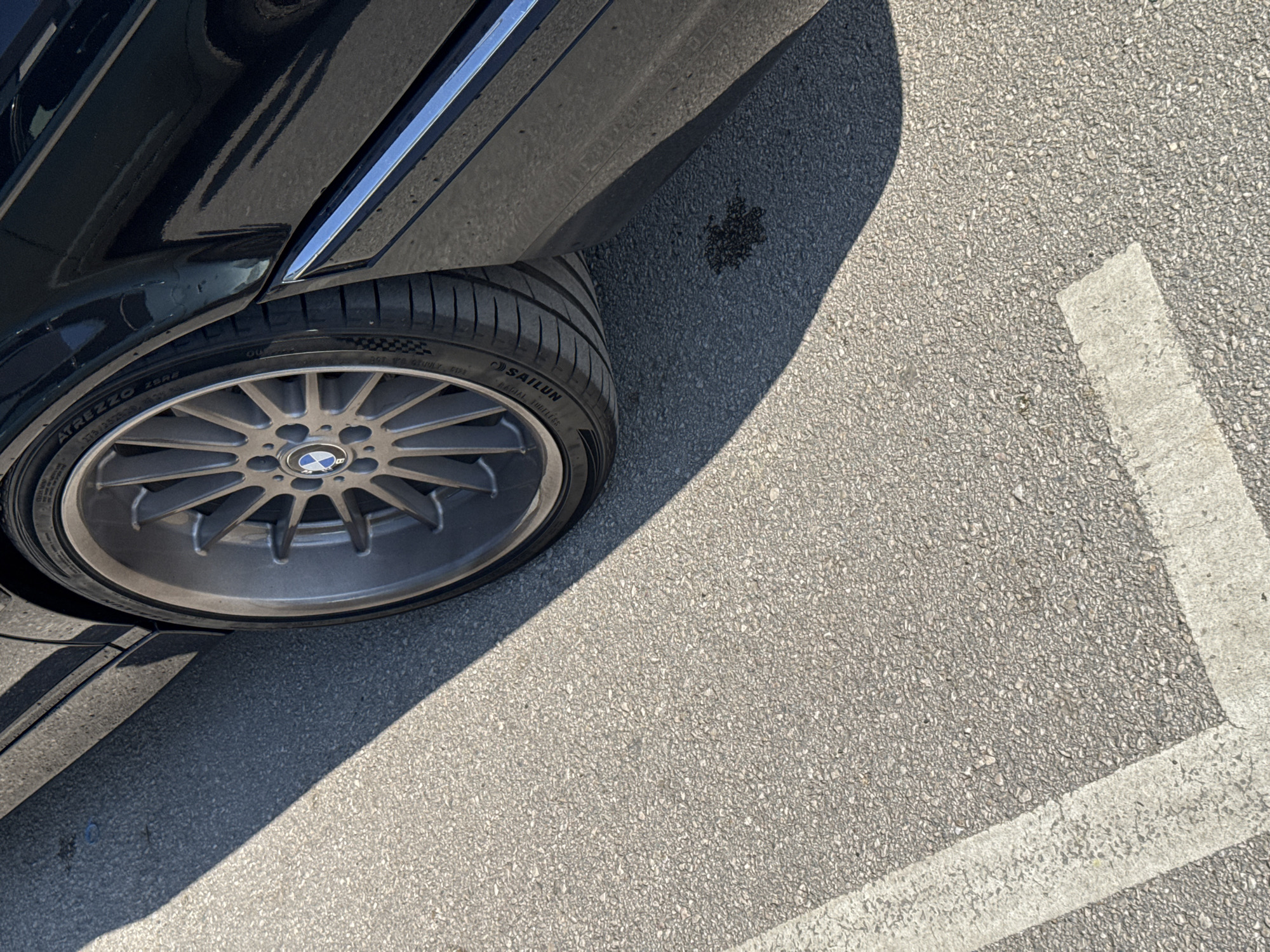 The image shows a close-up of the rear wheel of a black BMW car parked in a parking space. The tire is a Sailun brand. The parking space is marked with white lines on an asphalt surface. There is a small spot of liquid, possibly oil, on the ground near the wheel. The location is not evident from the image.