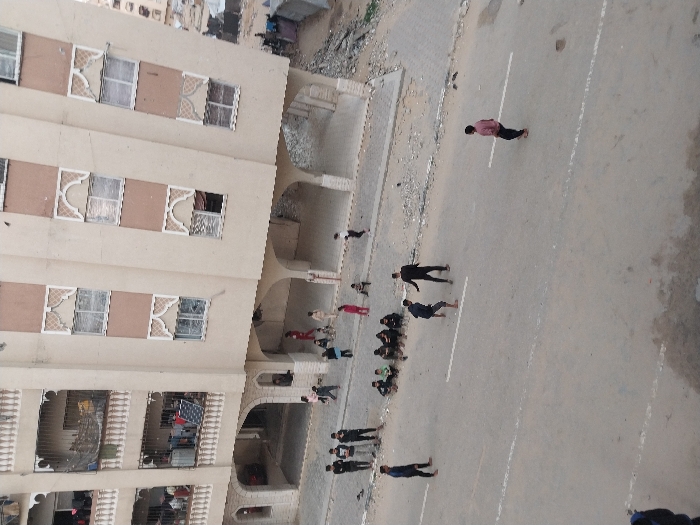 A group of people are gathered on a street next to a tall building. Some individuals are walking, while others appear to be sitting or standing in a line. The building has multiple floors with decorative windows and arched doorways. A white line runs along the center of the street, dividing the group from the more open pavement. The scene is shot from a high angle, overlooking the street and the building.