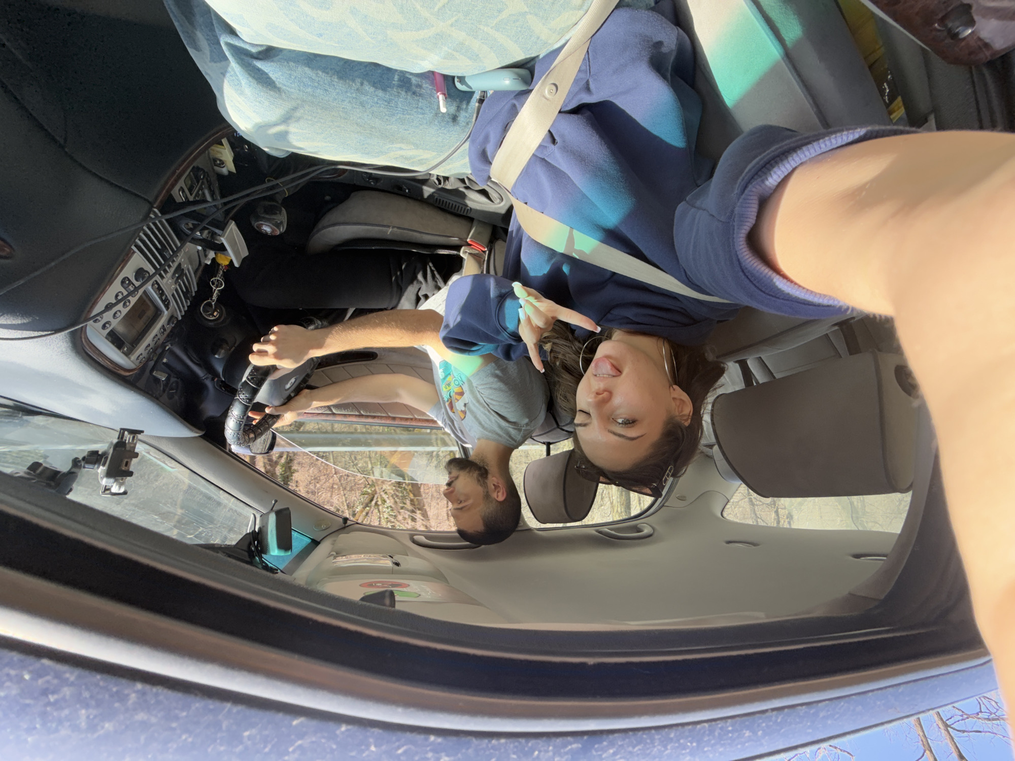The image shows the interior of a car, taken from the front passenger's perspective, with the camera facing towards the back. The interior is typical of a car with bucket seats in the front. The driver is a man with a beard. He is holding the steering wheel. The passenger, a young woman with dark hair, is making a peace sign with her fingers and sticking her tongue out. Both are wearing seatbelts. Outside the front windshield, bare trees and a blue sky are visible. A phone holder is visible on the windshield. Given the information provided ("location in the city of Sofia, Bulgaria"), this is an image that was taken in a car in Sofia.