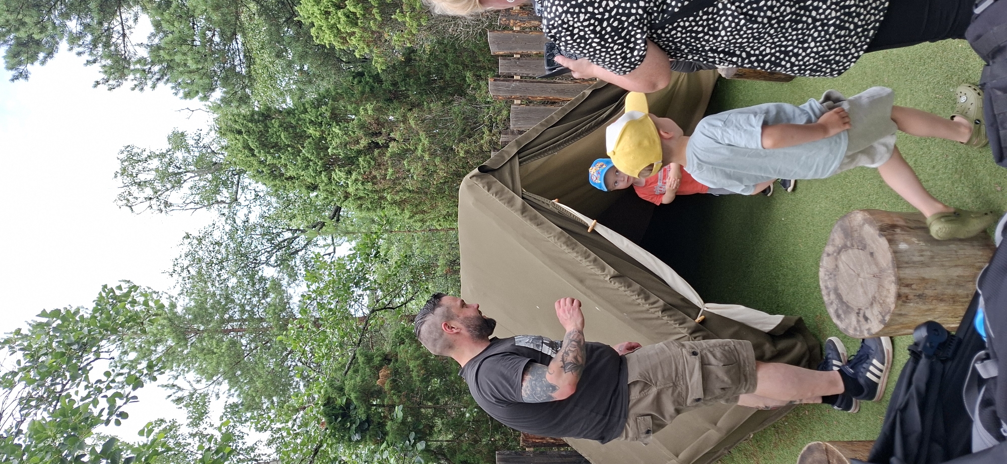 The image depicts an outdoor scene, possibly a park or a recreational area, with a focus on a canvas tent. In the foreground, a man with tattoos on his arm and a shaved head is wearing a dark t-shirt and khaki shorts. He appears to be interacting with something near the entrance of the tent. Two young children are visible near the tent. One child, wearing a red shirt and a blue cap with a logo, looks towards the camera from inside the tent's opening. Another child, in a light blue t-shirt and a yellow cap, is walking away from the tent. To the right, a person wearing a black and white patterned top is holding a smartphone, suggesting they might be taking photos or videos. The environment is characterized by lush green trees and a cloudy sky. Wooden log sections are placed on the ground, possibly serving as seating or decorative elements. The overall impression is a casual outdoor gathering or activity. Based on the context provided, this scene is located in the city of Naantali, Finland.