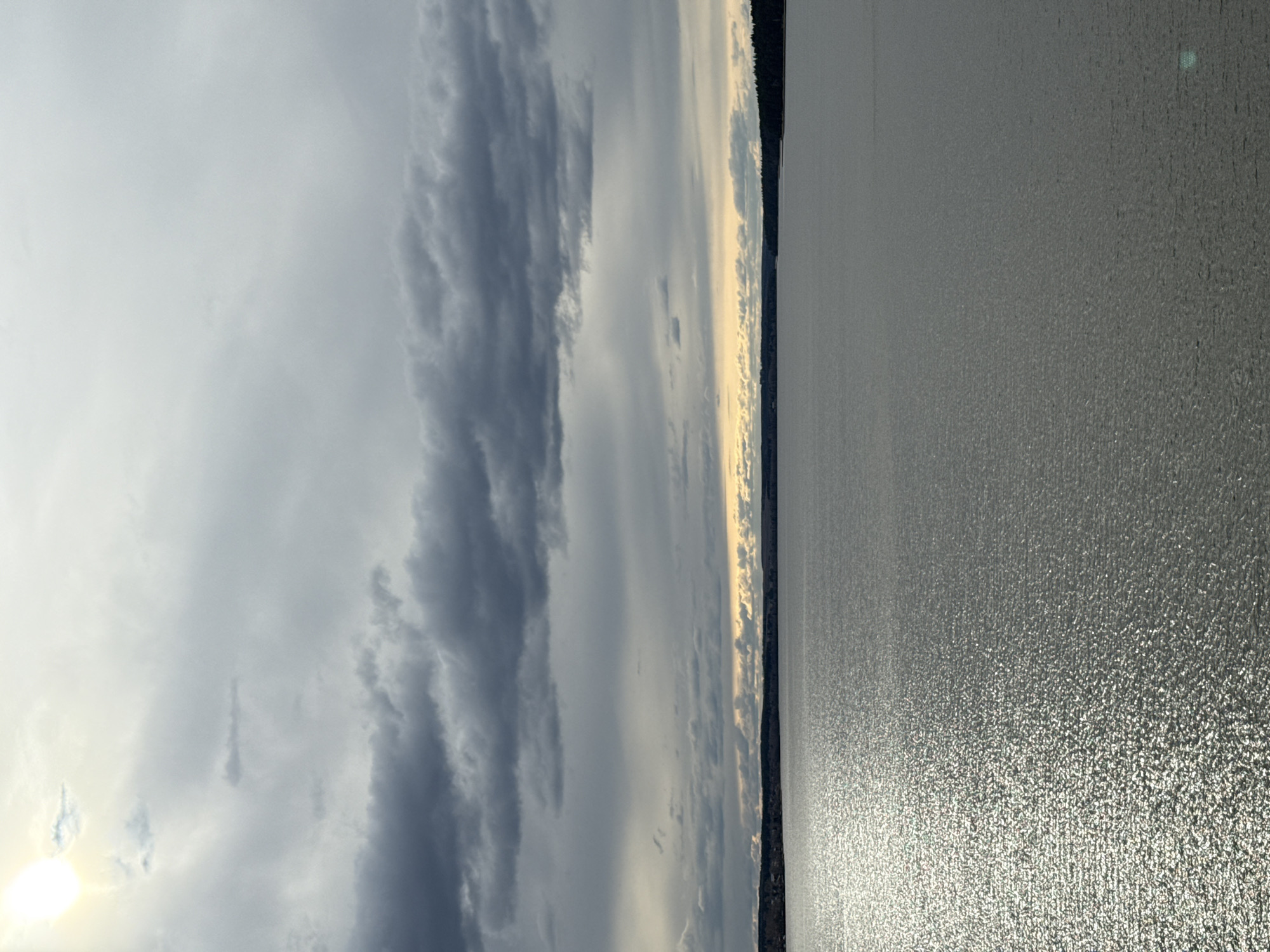 The image captures a vast outdoor landscape, predominantly featuring a sky filled with clouds and a large body of water, separated by a distant horizon. The scene is likely a lake or sea at either sunrise or sunset, given the low position of the sun.
The sky, occupying approximately two-thirds of the frame, is dramatic and dynamic. Dark, dense grey clouds stretch across the upper and right portions of the sky, creating a sense of depth and movement. Towards the left and central areas, the clouds are lighter and more diffused, allowing a bright sunburst to peek through on the far left.
A thin, dark line forms the distant horizon, suggesting a low-lying landmass or a dense treeline. Above this horizon, particularly towards the center, there are subtle golden and pale yellow hues in the clouds, illuminated by the sun.
The body of water fills the lower third of the image. Its surface is textured with countless sparkling reflections, especially prominent on the left where the sun's light directly hits the water, creating a shimmering effect. The water appears calm and vast.
There is no specific activity or event depicted, nor are there any people or artificial objects. The overall mood is one of tranquil grandeur and natural beauty, highlighting the interplay of light and clouds over a wide expanse of water. No text is visible in the image. The location is consistent with a large body of water, such as the Votkinsk Reservoir in Votkinsk, Russia.