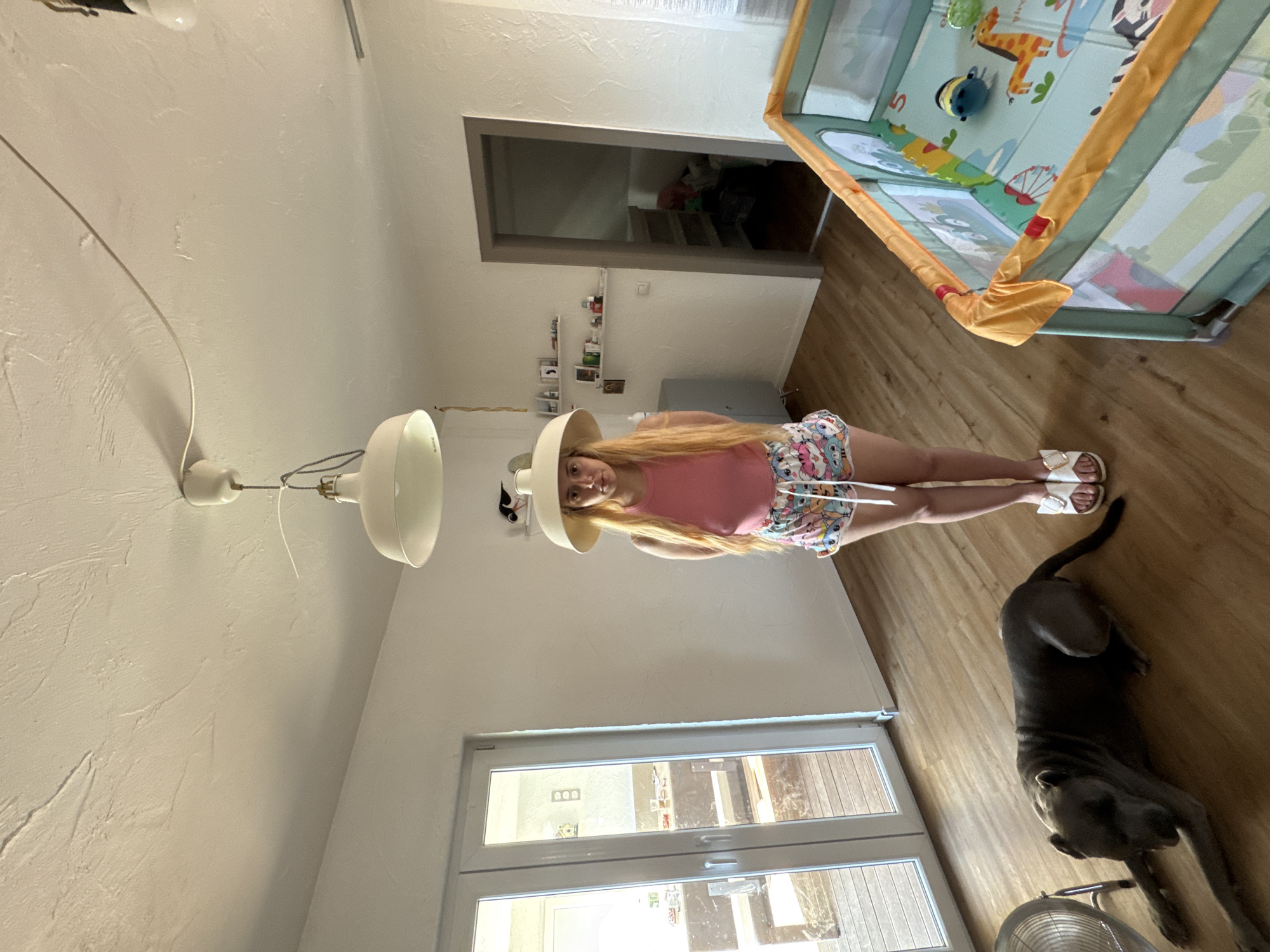 The media file shows a young woman with long blonde hair standing in what appears to be a living room. She is wearing a pink tank top and colorful patterned shorts, along with white slide sandals. A large, upside-down white lampshade is positioned over her head, obscuring part of her face. To her right, a dark-colored dog lies on the wooden floor. In the background, a baby playpen with colorful animal graphics is visible. The room has white walls and a wooden floor. A large window or glass door is also present on the left side of the frame, reflecting some of the interior. There is no indication of the time of day, weather, or specific location. The overall impression is a casual, indoor setting with an unusual photographic composition.