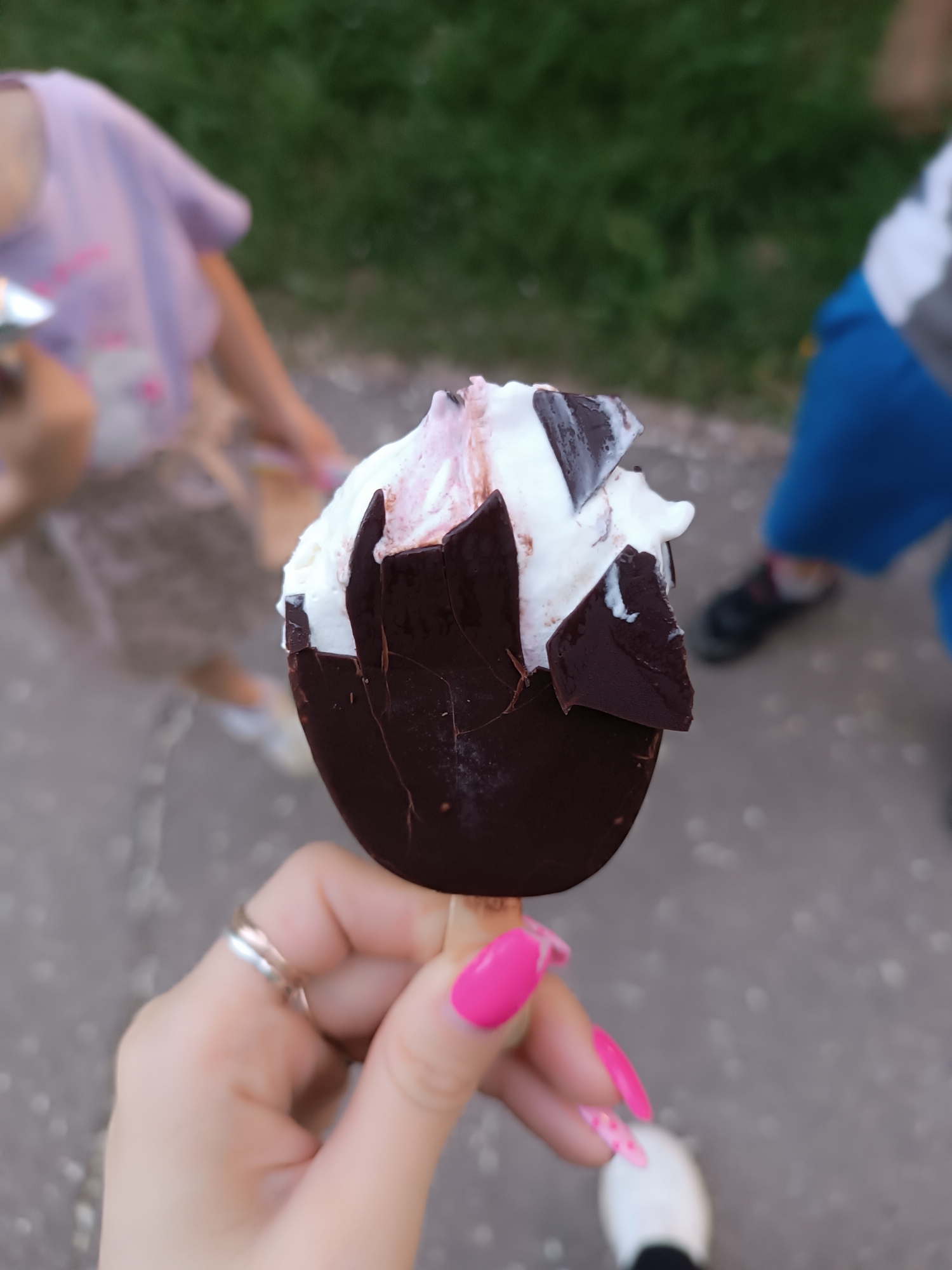 A hand with pink fingernails and a silver ring holds a partially eaten ice cream bar with a chocolate shell and vanilla and strawberry ice cream. The scene is an outdoor public space. Behind the ice cream bar are blurred figures, including a child wearing a pink shirt and skirt and another wearing blue shorts and black shoes, and a patch of green grass. The ground appears to be paved.