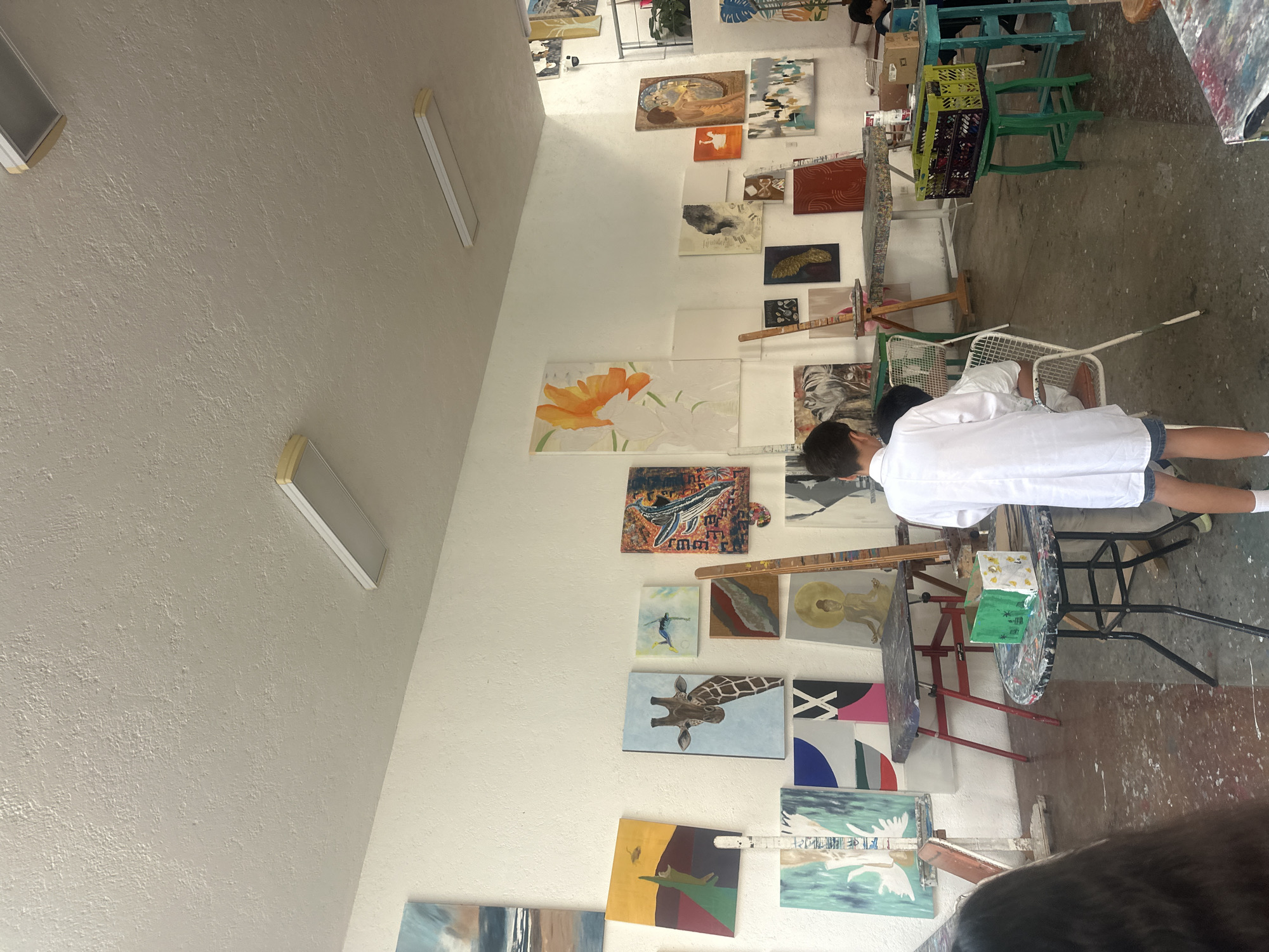 A white-walled room in San Bernardino Tlaxcalancingo, Mexico, contains numerous framed paintings displayed on the walls. A child in a white shirt is standing near a table, seemingly focused on an activity or object on it. Paint-splattered tables and chairs indicate an art studio or workshop environment. Light fixtures are visible on the high ceiling.