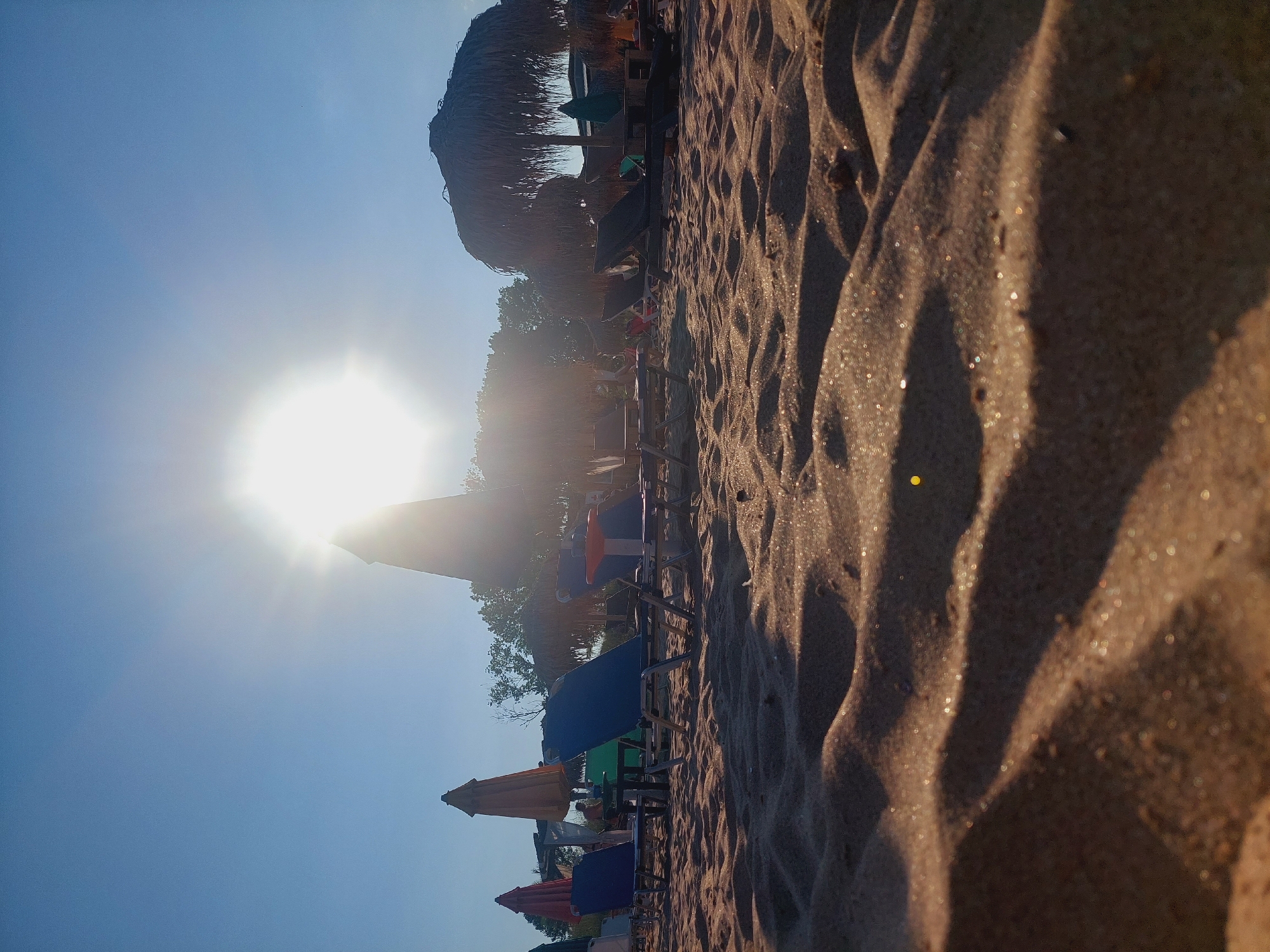 The image captures a sun-drenched beach scene in Sozopol, Bulgaria. The foreground is dominated by undulating sand dunes, with individual grains of sand glinting in the bright sunlight, creating a shimmering effect. In the midground, rows of empty beach chairs and umbrellas are arranged on the sand. Some of the umbrellas are made of straw, while others are fabric. Behind the chairs, a few trees are visible, their foliage silhouetted against the bright sky. The sun itself is positioned high in the sky, partially obscured by a tall, conical structure that could be part of a building or a large umbrella. The overall impression is one of a peaceful, early morning or late afternoon on a summer day.