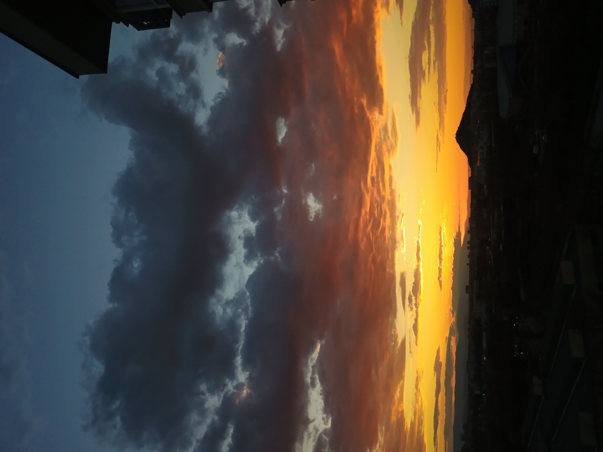 The image captures a sunset over Plovdiv, Bulgaria. The sky is a gradient of colors, transitioning from dark blue to orange and yellow, with clouds illuminated by the setting sun. The silhouette of a hill or mountain range is visible on the horizon. The photo was taken from a high vantage point, possibly a building, as parts of a building are visible at the edges of the frame.