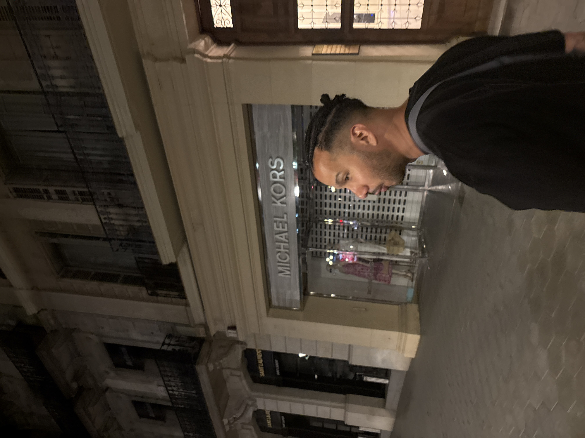 The photograph, taken at night in Barcelona, Spain, shows a man standing on a sidewalk in front of retail stores. The man is of African descent, with short, braided hair. He is wearing a dark-colored jacket or sweater. Behind him is a storefront with "MICHAEL KORS" emblazoned on the facade. Another storefront visible is "SAINT LAURENT." The buildings exhibit classic European architecture, featuring multiple stories, balconies, and decorative moldings. The scene is illuminated by streetlights and the glow emanating from the storefronts. The sidewalk appears to be paved with hexagonal tiles.