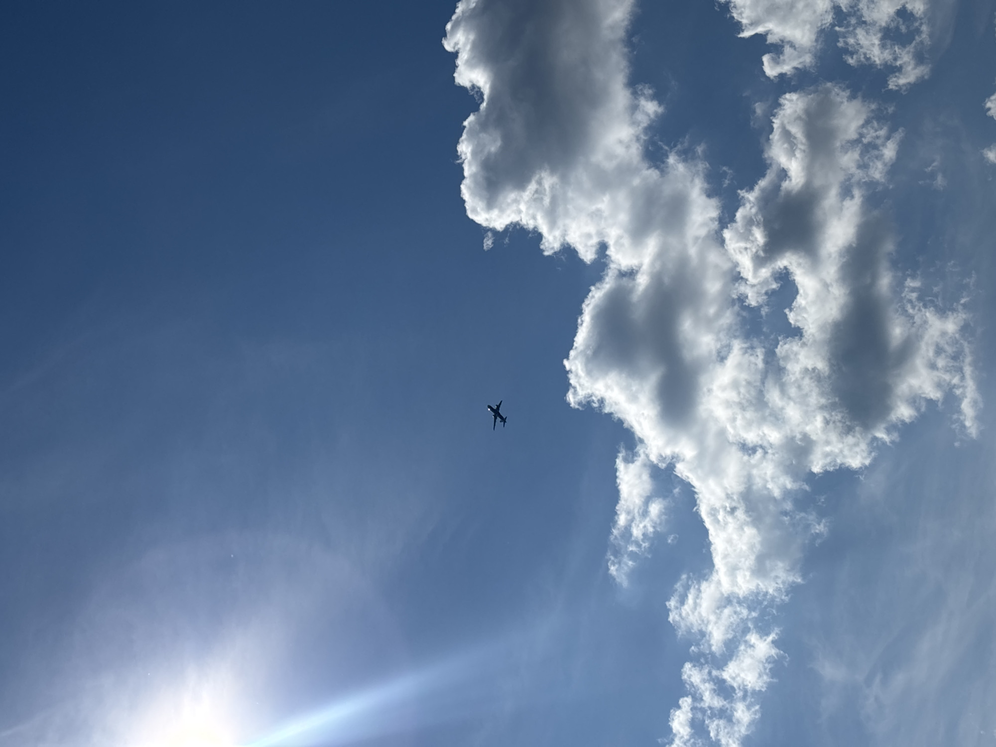 The media file depicts a bright, sunny day with a clear blue sky. A silhouetted airplane is captured in mid-flight, soaring against the vast expanse. Fluffy white clouds are scattered across the sky, with a prominent cluster of clouds occupying the upper right portion of the frame. In the lower left, a bright lens flare emanates from the sun, casting a subtle light across the scene. There is no visible text in the image. The overall impression is one of a peaceful daytime scene, possibly taken from ground level looking upwards. The location context of Novokuybyshevsk, Russia, suggests this is an outdoor scene in that region.