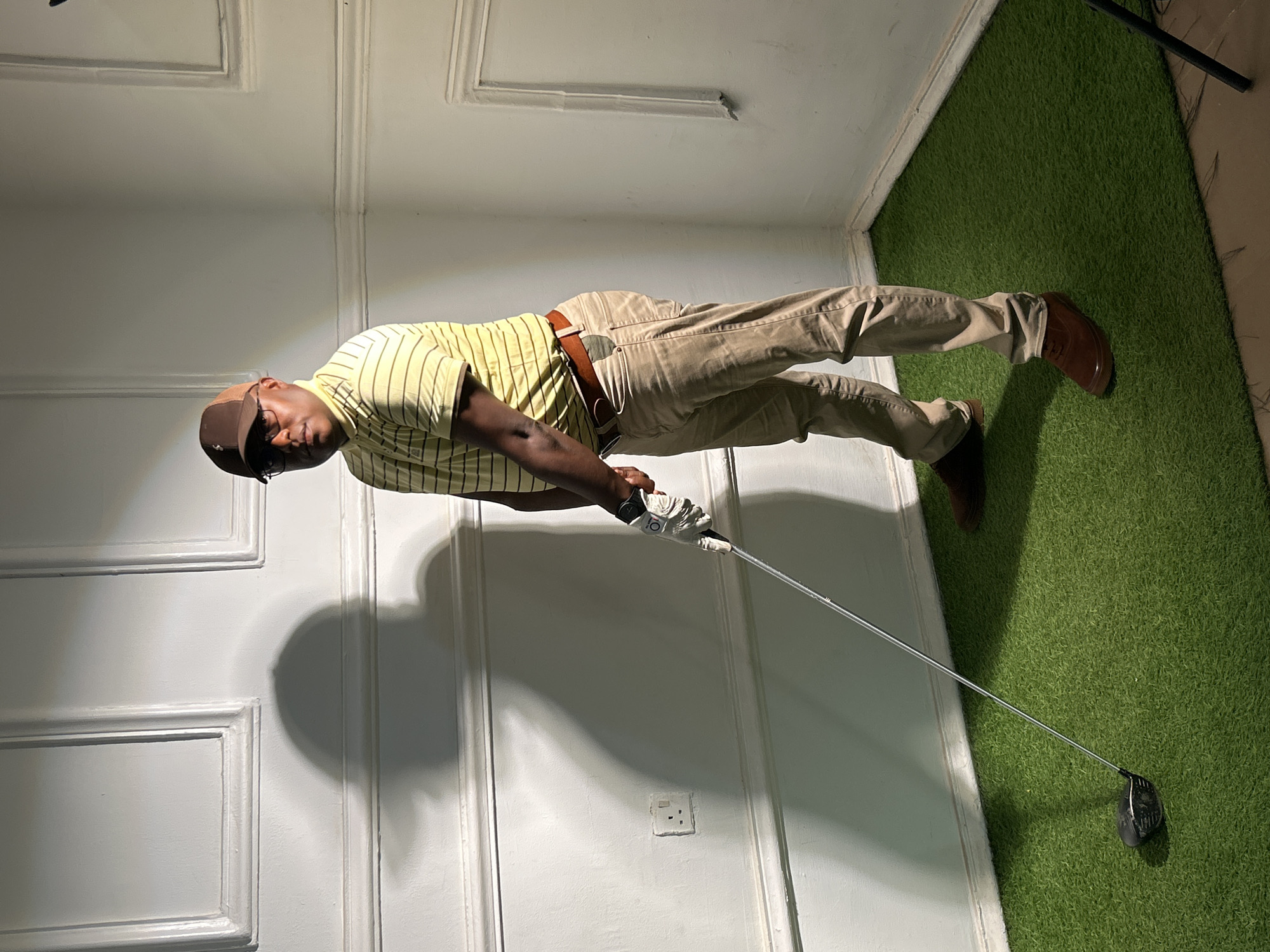 A man is standing indoors on a patch of artificial green turf. He is wearing a light yellow, short-sleeved, horizontally striped shirt, khaki pants held up by a brown leather belt, brown leather shoes, a brown baseball cap, and dark sunglasses. He is holding a golf club with a white glove on his left hand, appearing to be in mid-swing or about to swing.
The man's shadow is cast on a white wall behind him, which is panelled with a rectangular design and features a visible electrical outlet. The green turf appears to be an indoor setup, extending from the corner where the wall meets another wall. The upper part of a black tripod is barely visible in the right corner of the image.
There is no indication that this image was taken in Jos, Nigeria. There is no information in this image that suggests a specific location.