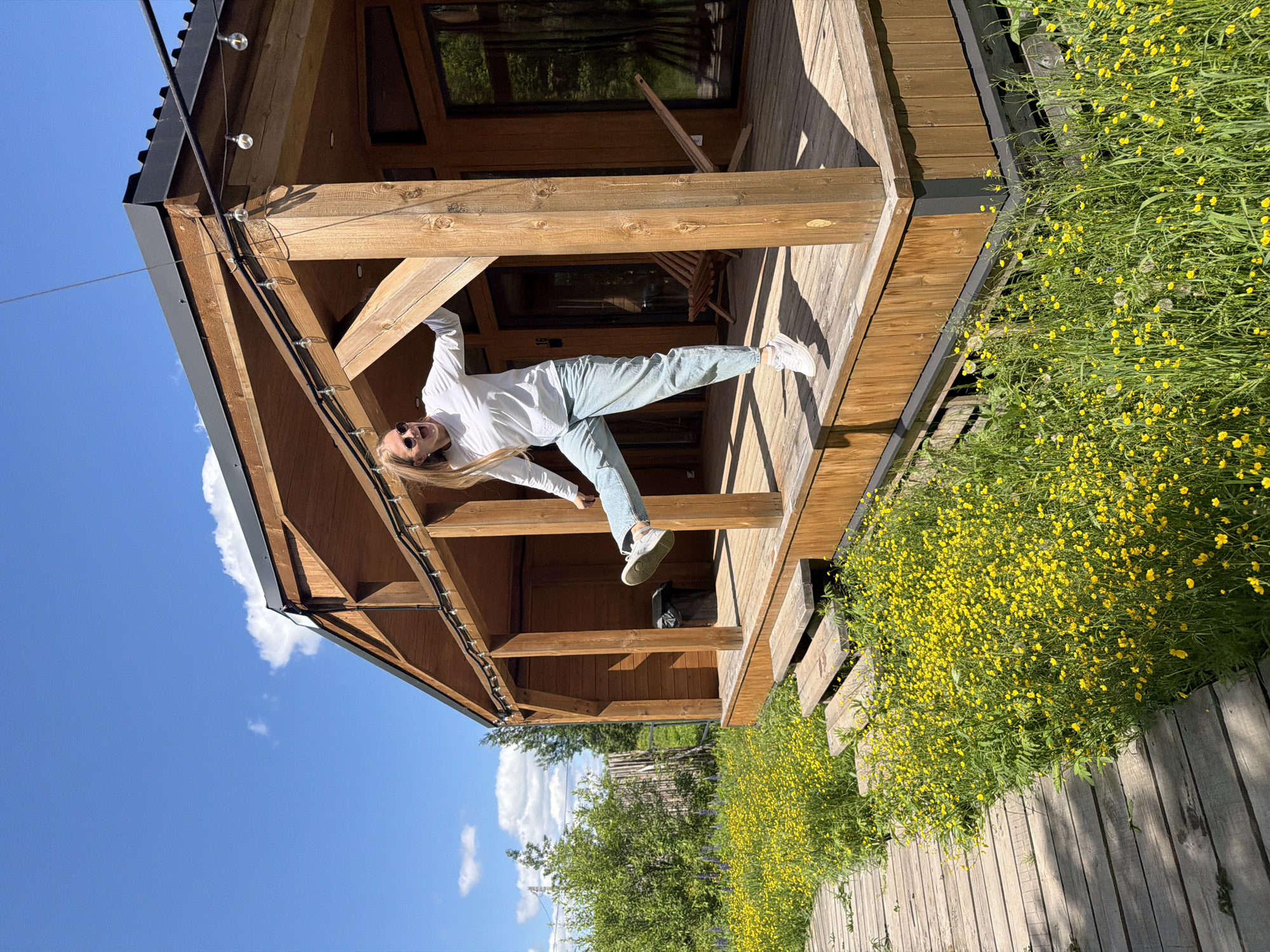 A vibrant outdoor photograph captures a young woman in a playful, acrobatic pose against the backdrop of a modern wooden cabin on a sunny day in Vozhoiskii, Russia.
The central subject is a woman with long blonde hair, dressed in a white long-sleeved top, light blue jeans, and white sneakers. She wears sunglasses and has a wide, open-mouthed smile, indicating joy and playfulness, as she suspends herself horizontally, holding onto a thick wooden beam of the cabin's porch. Her body is stretched out, suggesting a moment of fun and agility.
The setting features a well-constructed wooden cabin or house with a spacious porch, characterized by robust wooden beams, posts, and flooring. A dark roof, possibly metal, is visible, along with string lights (fairy lights) attached to the eaves of the porch. A window on the cabin reflects green foliage. The ground around the cabin is covered in lush green grass dotted with numerous small yellow flowers, resembling buttercups, creating a cheerful, natural landscape. A wooden plank pathway or boardwalk runs alongside this flowering area.
The sky is a clear, brilliant blue with a few fluffy white clouds, indicating bright, sunny weather during the day, likely late morning or early afternoon, as evidenced by the distinct shadows cast by the wooden structures. There is no visible text in the image. The overall scene conveys an atmosphere of joyful outdoor leisure in a scenic, natural environment.