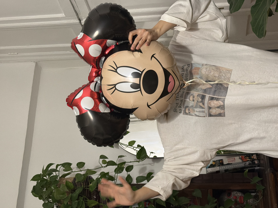An individual stands indoors, their face completely obscured by a large, foil Minnie Mouse head-shaped balloon featuring black ears, a red polka-dotted bow, and a smiling facial graphic. The person wears a loose-fitting, light-colored long-sleeved t-shirt with a graphic print displaying multiple figures and partial text, including "Brad: The... to look like h... girlfriend". Their right hand (viewer's left) is raised, palm open, as if in a wave, while their left hand (viewer's right) holds the balloon. In the background to the left, a potted plant with green trailing vines resides in a woven basket, situated on dark wooden furniture. Below the plant, several books or media cases, one with a faint "Marvel" logo, are visible. A portion of a mirror reflects the room behind the plant. The background wall is plain and light-colored, with a white doorframe visible on the right. This scene takes place indoors in the city of Moscow, Russia.