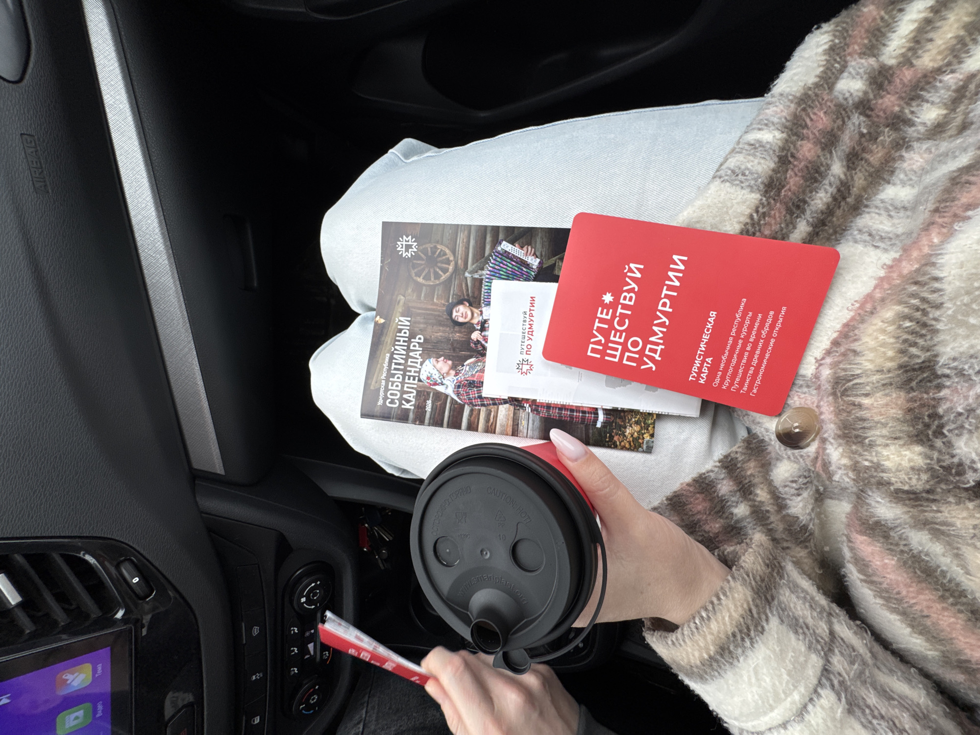Here is a description of the image:
**Scene:** The image appears to be taken inside a car, likely in Votkinsk, Russia, based on the Cyrillic text on the documents.
**People:** A person is visible by their hands and a portion of their arm wearing a patterned coat. Their hands hold a coffee cup with a black lid and appear to be adjusting something on the car's dashboard. The person has light skin and manicured nails.
**Subjects:** The main subjects are promotional tourism documents related to the Udmurt Republic in Russia, and a cup of coffee.
**Objects:**
* A red tourism brochure titled "Путешествуй по Удмуртии" (Travel in Udmurtia).
* A calendar related to events in the Udmurt Republic.
* A coffee cup with a black lid.
* A car dashboard with buttons, controls, and a display screen.
* The car's interior, including the airbag panel and part of the seat.
**Actions/Interactions:** The person is holding the coffee cup and appears to be interacting with the car's dashboard, possibly with a red-and-white stick-like object. The tourism brochures and calendar are resting on the person's lap.
**Environment:** The environment is the interior of a car. The light indicates it is possibly daytime, but the darkness inside the car suggests either overcast weather or that the image was taken in shadow.