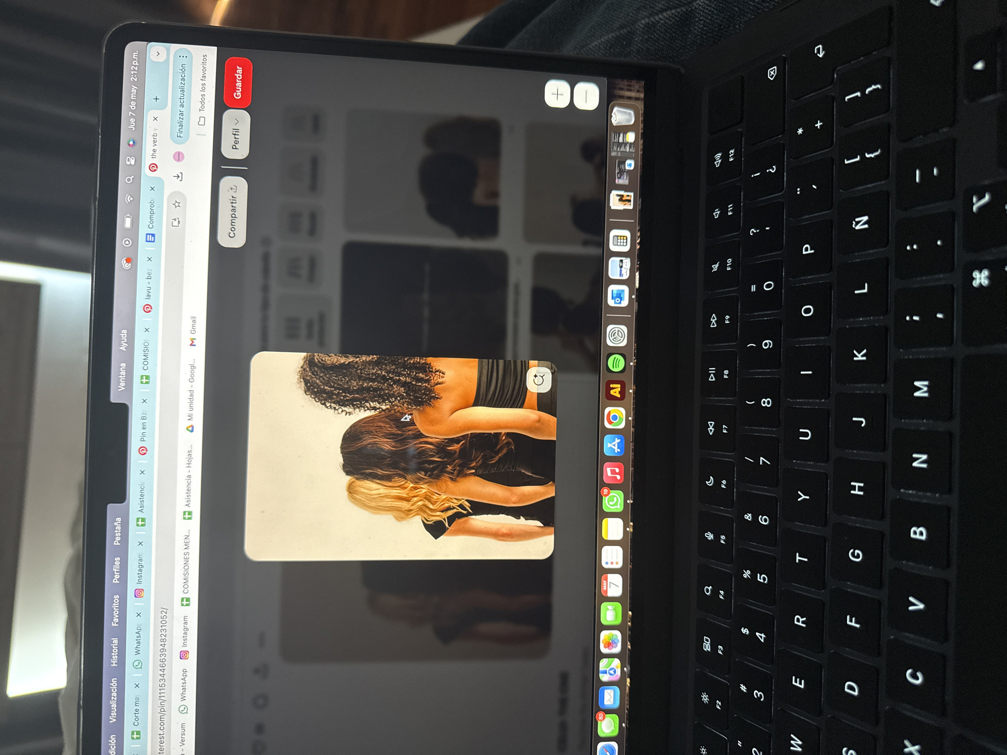 A close-up shot shows a black MacBook open, displaying the Pinterest website. On the right side of the screen is the keyboard, which is black with white lettering. The screen displays multiple open tabs, including WhatsApp, Instagram, and Gmail. The main focus of the screen is the Pinterest tab, displaying an image of three women in black dresses, with hair in three different shades: blond, brown, and black. On the right of the screen, there is a column of app icons, including Adobe Illustrator, Google Chrome, and WhatsApp. The date and time "Jue 7 de may 2:12 p.m." are visible in the upper left corner of the screen. The location of the scene is not discernable from the image.