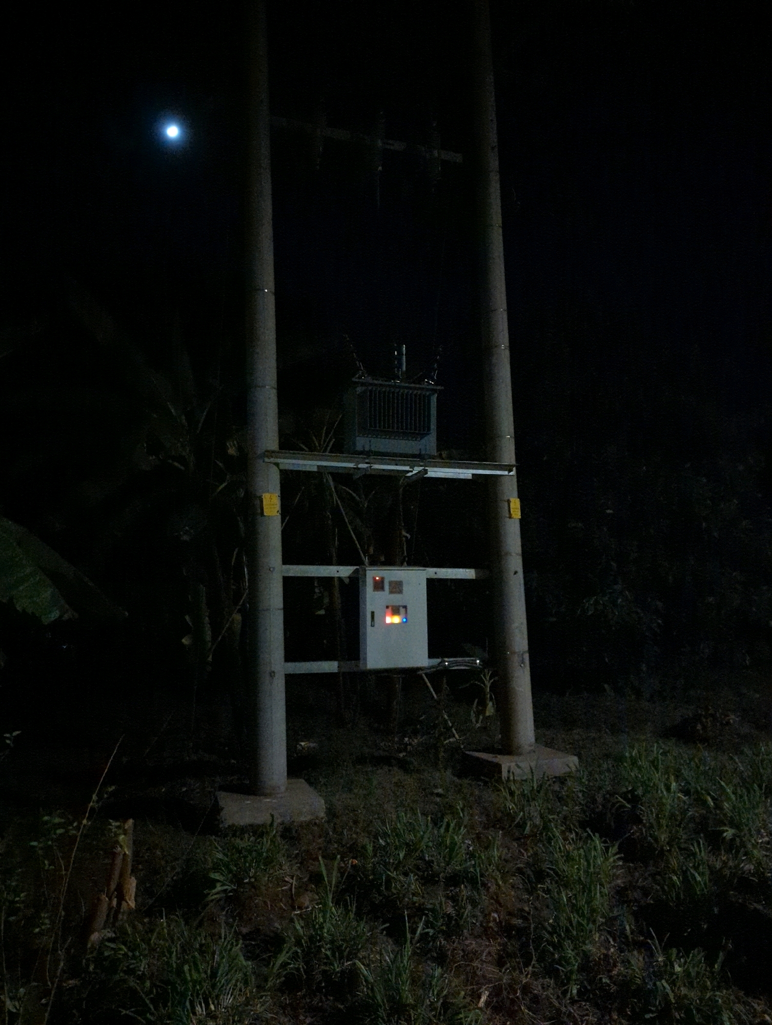 The image depicts an outdoor scene at night, presumably in the Musanze District of Rwanda. The primary focus is a utility pole with electrical equipment mounted on it. Two tall, cylindrical concrete poles support a transformer and a control box. The transformer, a dark-colored, rectangular apparatus with visible fins, is positioned higher up on the poles. Below it, a lighter-colored control box contains what appear to be indicator lights, one of which is illuminated in red and yellow. A bright, circular object, likely the moon, is visible in the dark sky to the upper left of the scene. The poles are situated in a grassy area with some vegetation visible in the background and foreground, suggesting a rural or semi-rural setting. The overall lighting is dim, with the electrical equipment and the moon being the main sources of illumination.