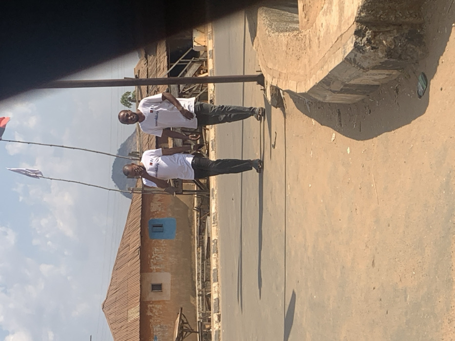 In Egbedi, Nigeria, two adult males walk on a paved road under a clear, sunlit sky. Both individuals wear identical white short-sleeved t-shirts with a dark emblem on the chest, and dark-colored trousers. The male on the left, with short dark hair and a beard, holds a dark rectangular object to his right ear, with his mouth slightly open, suggesting communication. He wears dark open-toed footwear. The male on the right, bald and clean-shaven, looks forward and slightly to the right of the viewer, wearing light-colored open-toed footwear. Long shadows are cast from their figures to the left on the road surface. In the background, a single-story building with a reddish-brown corrugated metal roof and light-brown walls is visible, featuring a prominent blue-painted window. Behind it, a conical hill rises, flanked by two tall bamboo poles, each carrying a flag; the left flag is white, the right is red and dark. The immediate foreground on the right consists of a dusty dirt shoulder bordering a concrete curb, partially obscured by a dark vertical post.