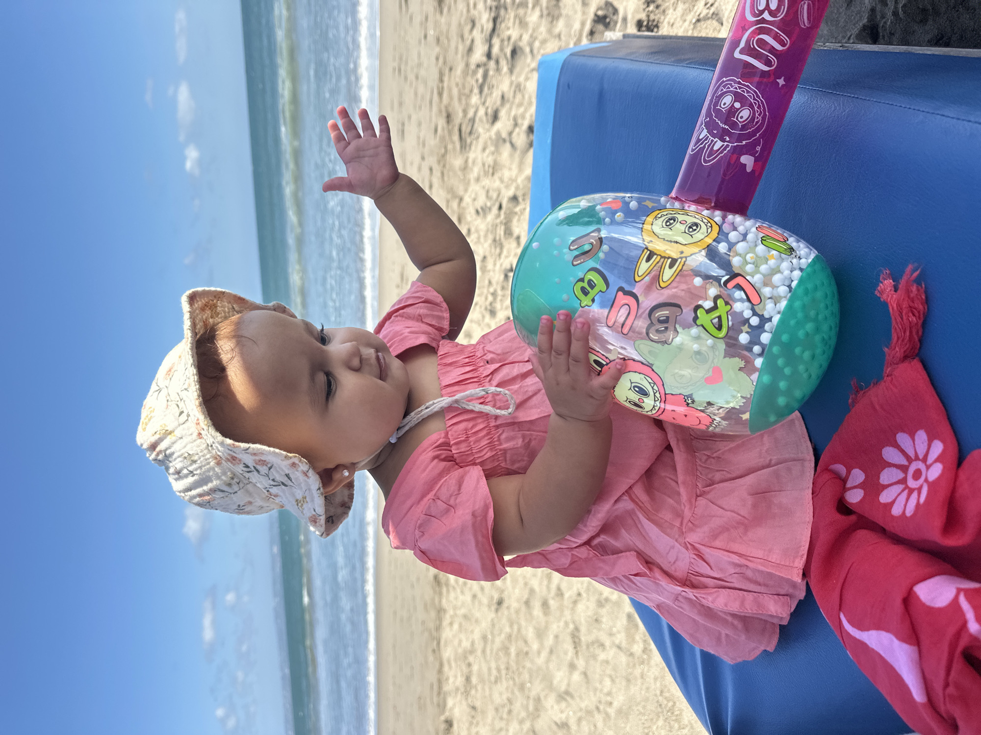 A baby girl wearing a sun hat and a pink dress sits on a blue beach chair on a sandy beach. The ocean with gentle waves is in the background. The baby is holding a colorful inflatable toy. Her right hand is raised, as if waving. The toy appears to have cartoon characters and letters printed on it, and contains small white beads. A pink towel with a floral pattern is draped over the chair. The image captures a sunny day, likely mid-morning or afternoon, given the bright lighting and clear blue sky. The setting is a beach in North Kuta, Indonesia. The baby appears content and engaged with her toy. There is some text visible on the inflatable toy, including letters and what appear to be cartoon characters.