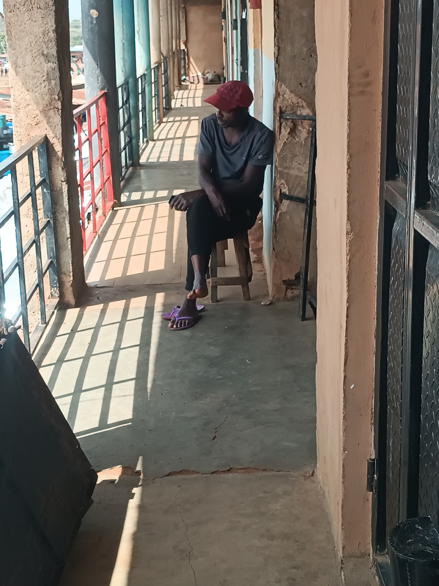 A person wearing a red cap, gray shirt, and dark pants is sitting on a wooden stool in a covered walkway. The walkway is lit by sunlight creating sharp shadows from a metal railing along one side. The railing has red and gray vertical supports. The walkway is part of a building with crumbling walls and painted pillars. The person is facing away from the railing. One foot is resting on a purple sandal. This scene is located in Bukuru, Nigeria.