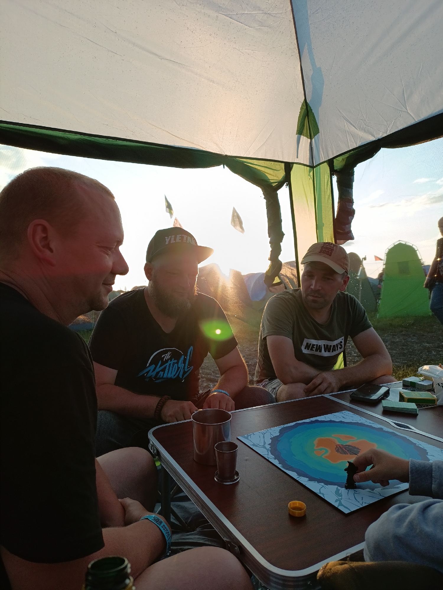 Three men are sitting at a folding table under a canopy, possibly at a festival or campsite, with tents and flags visible in the background. The man on the left, facing away from the camera, has a shaved head and is wearing a black t-shirt. The man in the center, wearing a black t-shirt with a graphic and a baseball cap, has a beard and is looking down. The man on the right, wearing a tan baseball cap and a green t-shirt with "NEW WAYS" printed on it, is looking forward. On the table, there is a board game with colorful concentric circles, a game piece, several metal cups, a smartphone, and some boxes. The sun is setting in the background, casting a warm glow and creating lens flare. The canopy shows signs of condensation or rain. The location is stated to be Lagunovo, Russia.