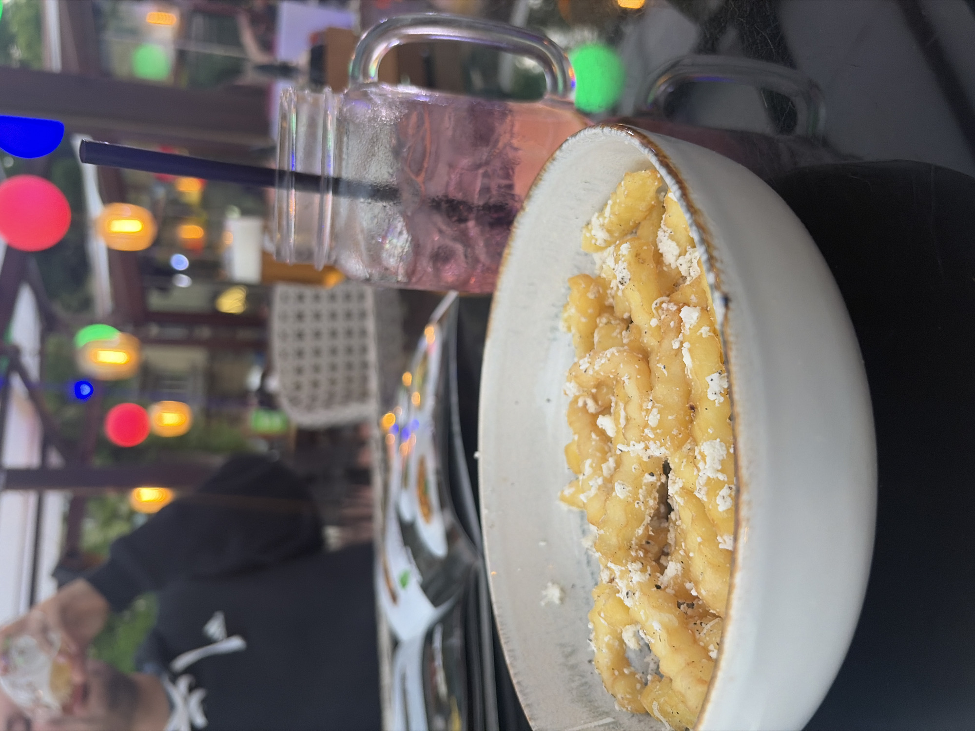 The image, rotated 90 degrees counter-clockwise, captures a casual dining setting, possibly in Dobrich, Bulgaria. In the foreground, a white bowl with a rustic brown rim contains a serving of food resembling pasta or gnocchi, characterized by twisted, tubular shapes. It is generously topped with grated white cheese, likely Parmesan or feta, and sprinkled with black pepper.
Beside the food is a clear, jar-style glass mug with a handle, filled with a pink or purple cold beverage. A black straw is inserted into the drink, and condensation is visible on the outside of the glass.
Partially visible in the lower left of the frame (due to the rotation) is a person wearing a black hoodie that features a white logo, possibly the Adidas symbol. The person, who has a beard, appears to be holding an object in their hand.
The background is softly blurred with numerous colorful bokeh lights in blue, red, orange, and green hues, suggesting an evening or nighttime atmosphere. Blurry architectural elements, including what appears to be a lattice pattern and structural beams, are also present. The scene conveys a relaxed and enjoyable dining experience. No other discernible text is visible apart from the potential logo on the hoodie.