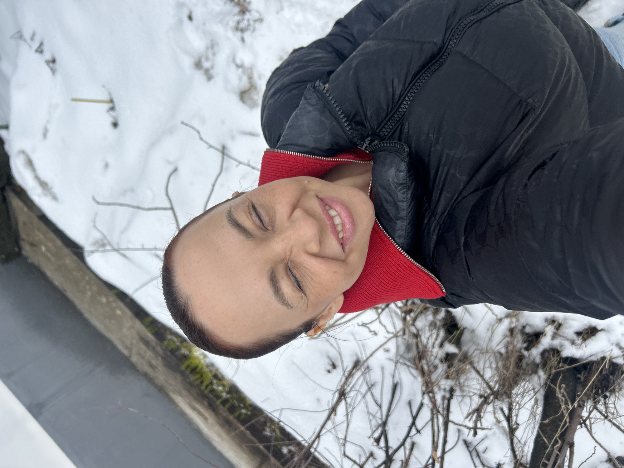 A woman is taking a selfie in a snowy outdoor setting in what appears to be the city of Chaikovsky, Russia. The woman is fair-skinned, with her hair pulled back, wearing a black puffer jacket with a red collar. She is smiling with her eyes closed. The background includes snow-covered ground, small branches, and a concrete structure covered with green moss.