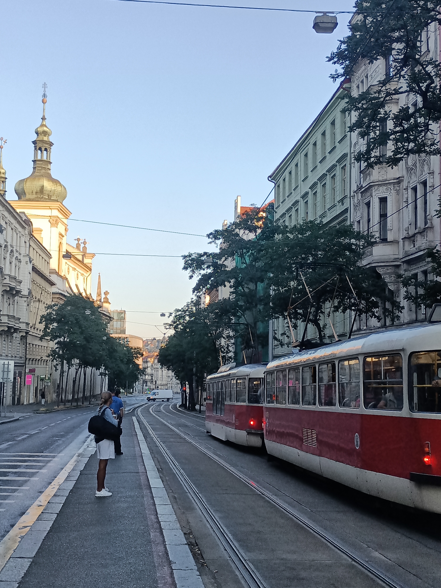 The image depicts a street scene in Prague, Czechia, likely during the late afternoon or early evening based on the lighting. Two red and white trams are parked on parallel tracks on the right side of the street. The tram on the right is closer to the viewer, showcasing its windowed interior. On the left side of the street, a person with blonde hair and white shorts stands on a sidewalk, looking towards the street. Behind them, another person is visible in blue. The street is paved and wet, suggesting recent rain. Buildings with traditional European architecture line both sides of the street, with a prominent church tower with a golden dome visible on the left. Trees are planted along the sidewalks. A white van is seen driving in the distance down the street. The sky is clear and pale blue.