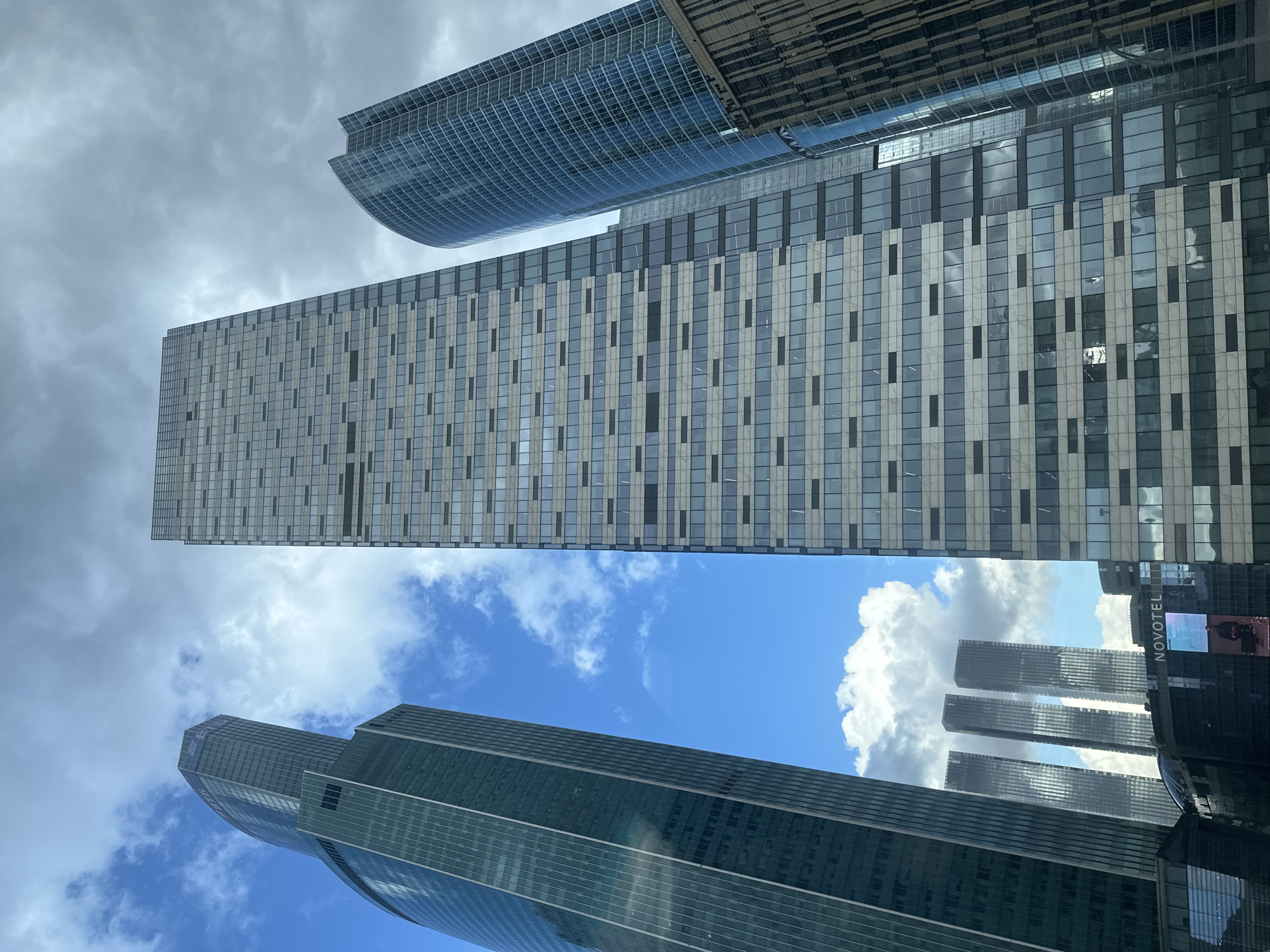 The image captures a low-angle view of several skyscrapers in the Moscow International Business Center (Moscow City), Russia. The buildings are predominantly glass and steel structures with a modern architectural style. The sky is visible between the buildings, with patches of blue sky interspersed with white, puffy clouds. One building has a distinct, curved shape at the top. Another has a pattern of alternating vertical bands of light and dark panels. The "NOVOTEL" hotel sign is visible on the side of one of the buildings.