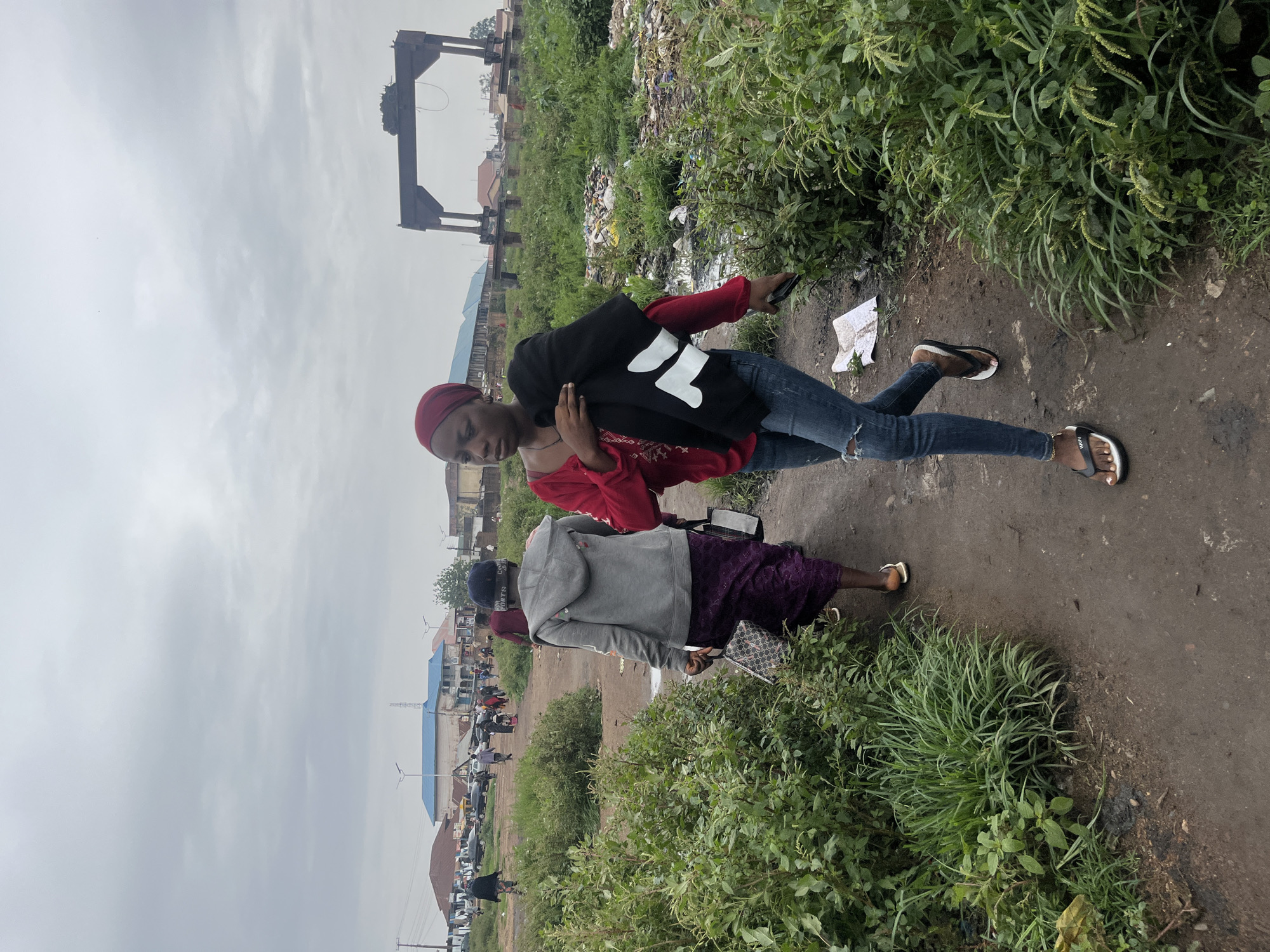 In Bukuru, Nigeria, under an overcast sky, two women walk on a dirt path lined with lush green vegetation and scattered debris. The woman in the foreground, wearing a red top, ripped blue jeans, and flip-flops, carries a black garment over her right arm. To her left, another woman walks away from the camera, dressed in a grey hooded jacket and a purple patterned skirt, carrying two bags. In the background, a large industrial structure, resembling a gantry crane, stands against the sky. Further in the distance, buildings and more people are visible, suggesting a populated area. The ground is a mixture of dirt and patches of water, with some areas heavily littered with plastic waste.