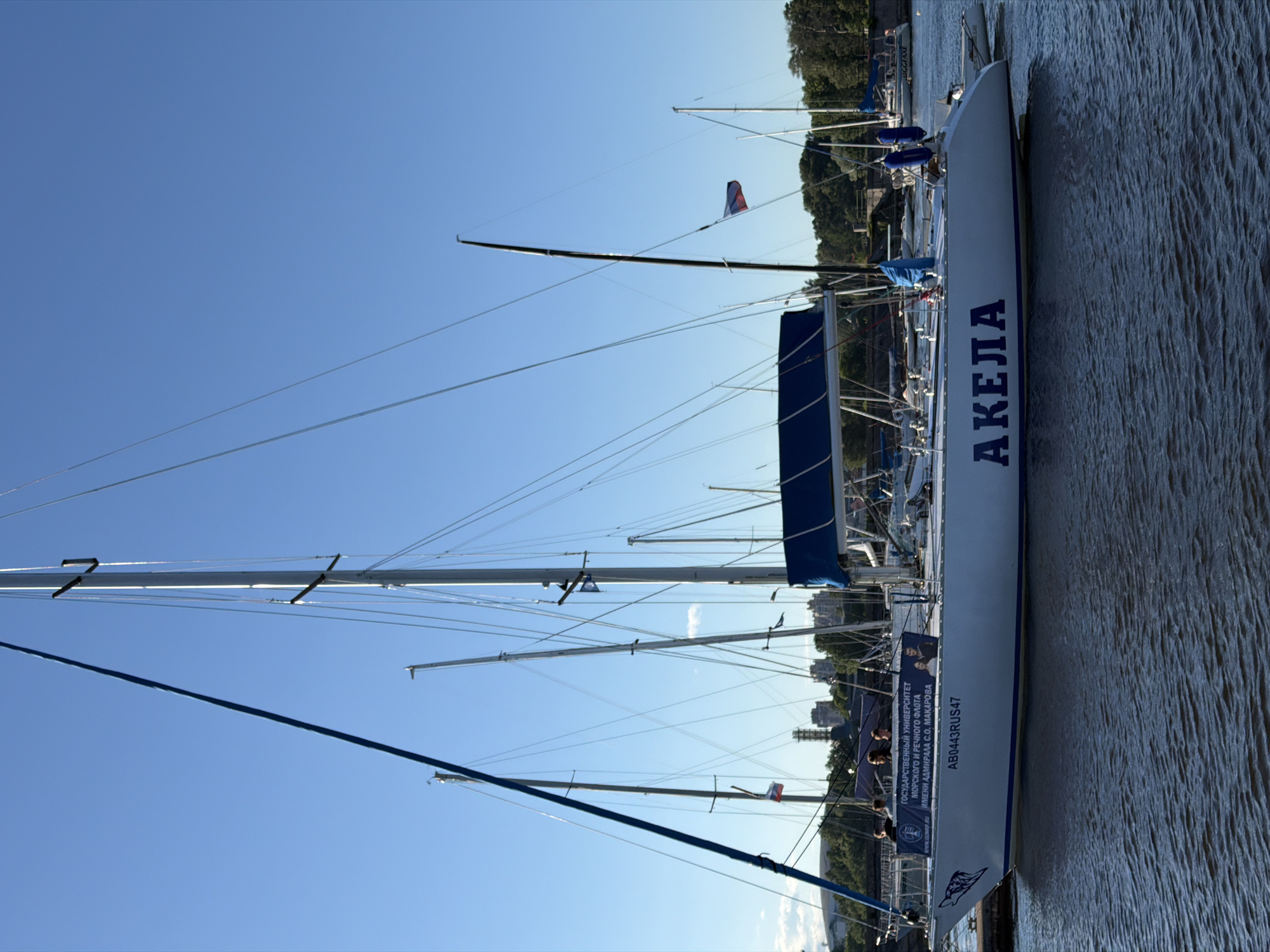 A white sailboat named "АКЕЛА" is docked on the Neva River in Saint Petersburg, Russia. The sailboat has a blue stripe along its hull and a blue canopy over its deck. On the side of the boat, there is a banner with text in Russian that reads "ГОСУДАРСТВЕННЫЙ УНИВЕРСИТЕТ МОРСКОГО И РЕЧНОГО ФЛОТА ИМЕНИ АДМИРАЛА С.О. МАКАРОВА" and "AB0443RUS47". Several people are visible on the deck of the sailboat, and a Russian flag is flying from one of the masts. The background shows a clear blue sky, trees, buildings, and other sailboats.