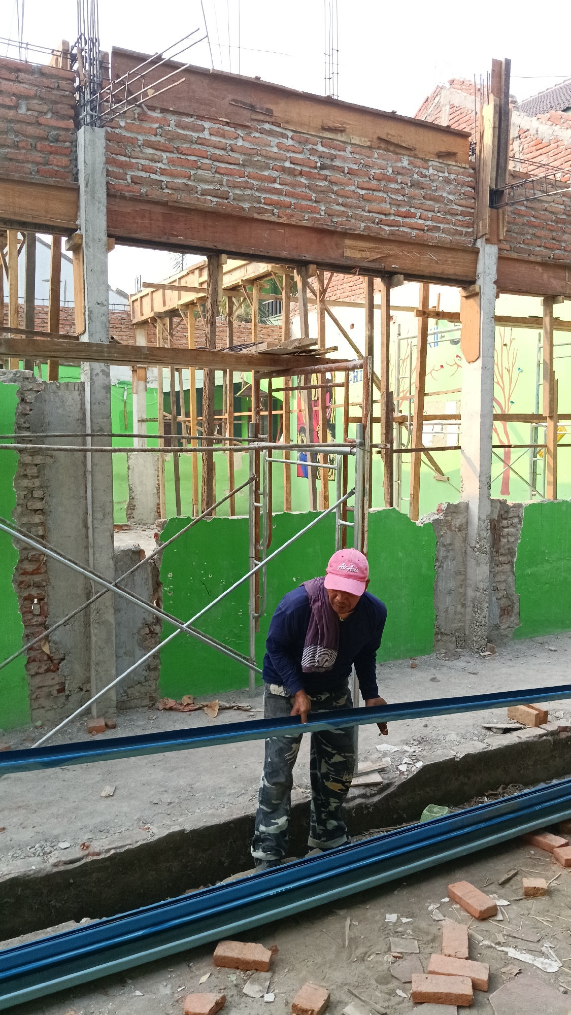 In Branggahan, Indonesia, a construction worker wearing a pink baseball cap, a blue long-sleeved shirt, and camouflage pants is seen on a construction site. The worker is bent over, holding a blue metal beam. The background shows an unfinished building with exposed brickwork, wooden beams, and green painted walls. Scaffolding and construction materials like bricks and metal rods are visible. The ground is dusty and littered with debris.